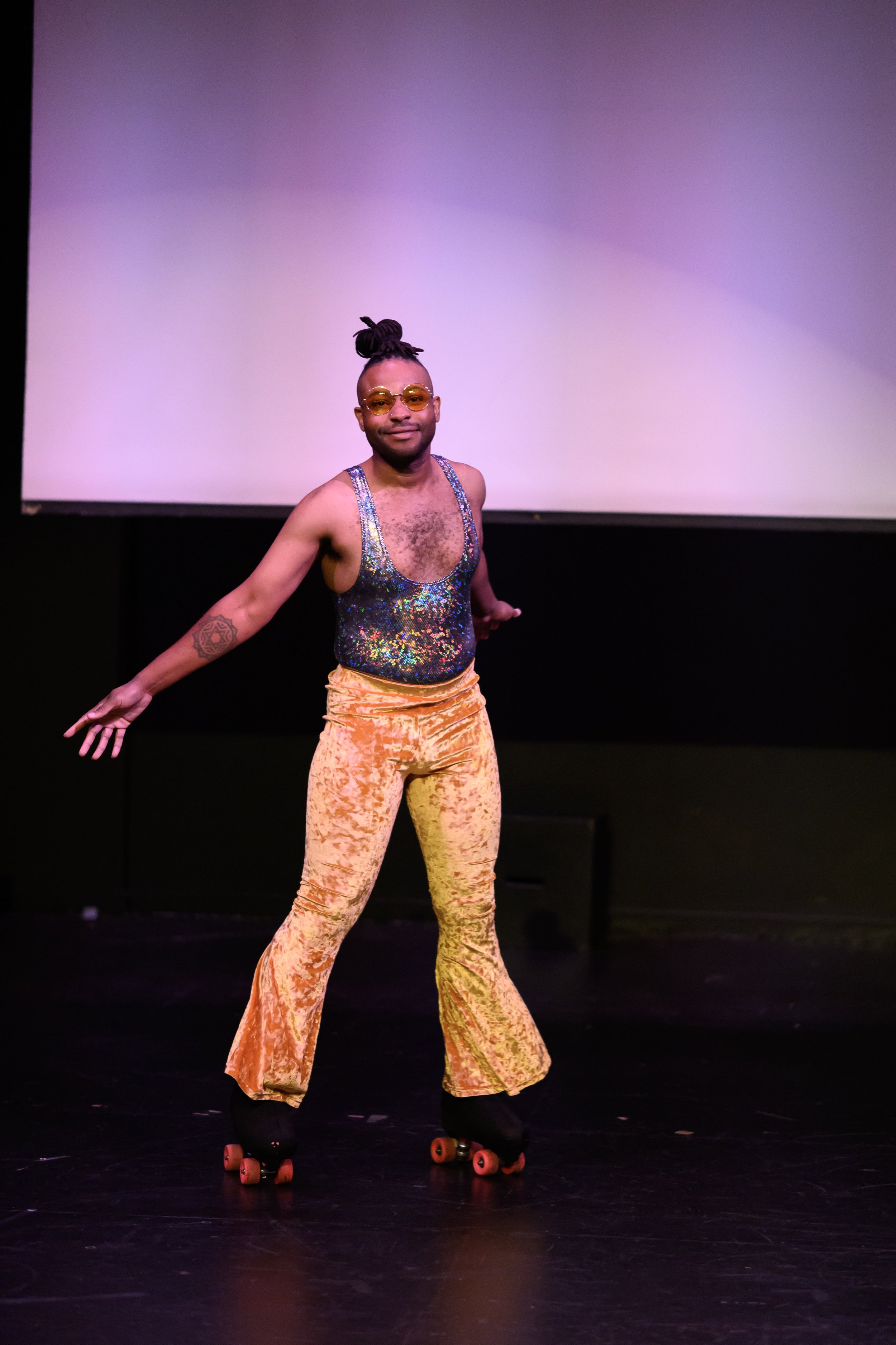Person in shiny tank top and velvet bell-bottom pants roller skating on stage