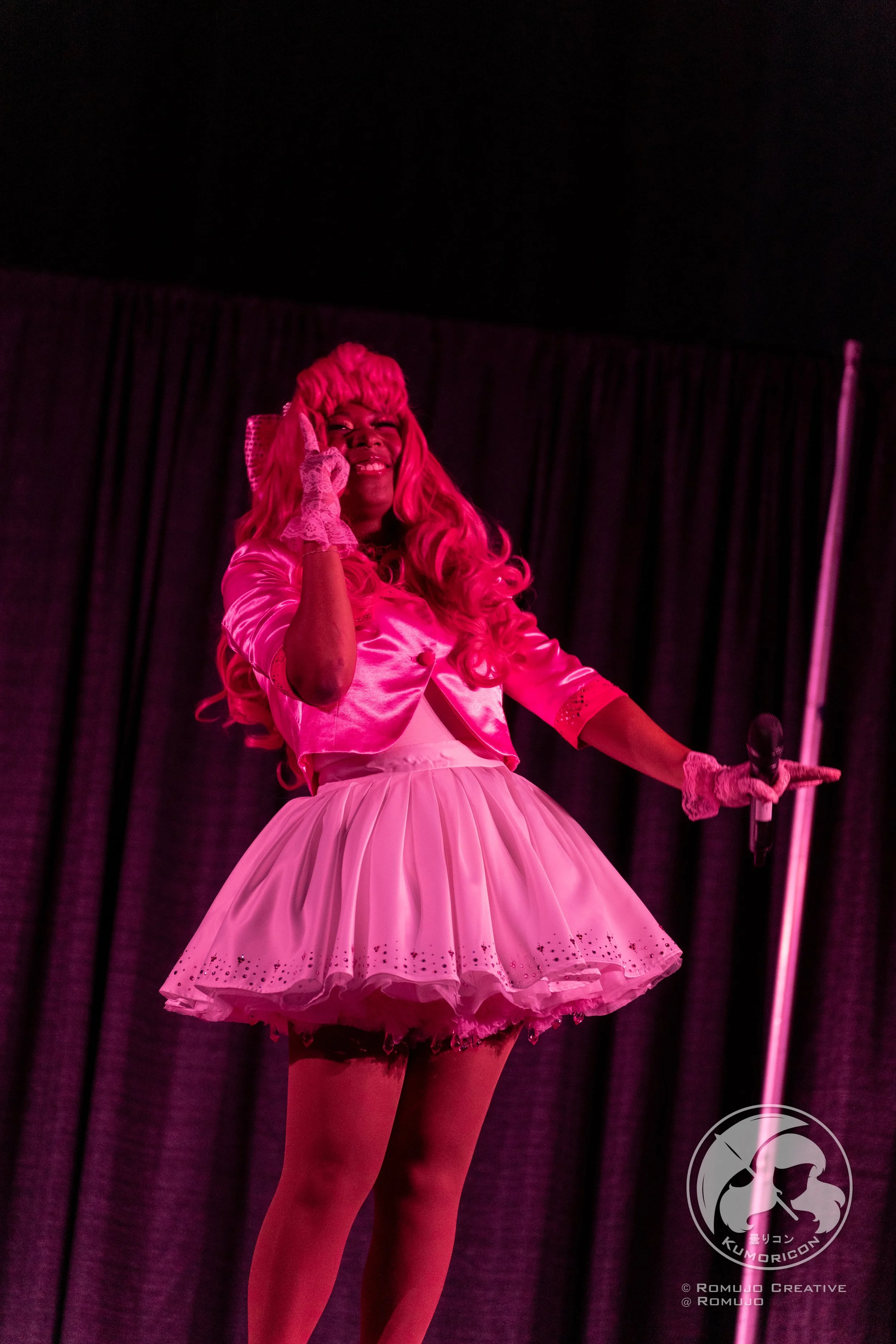Performer dressed in pink with long wavy pink hair, wearing a satin jacket and a short frilly skirt, on stage with a black curtain background, holding a microphone, smiling.