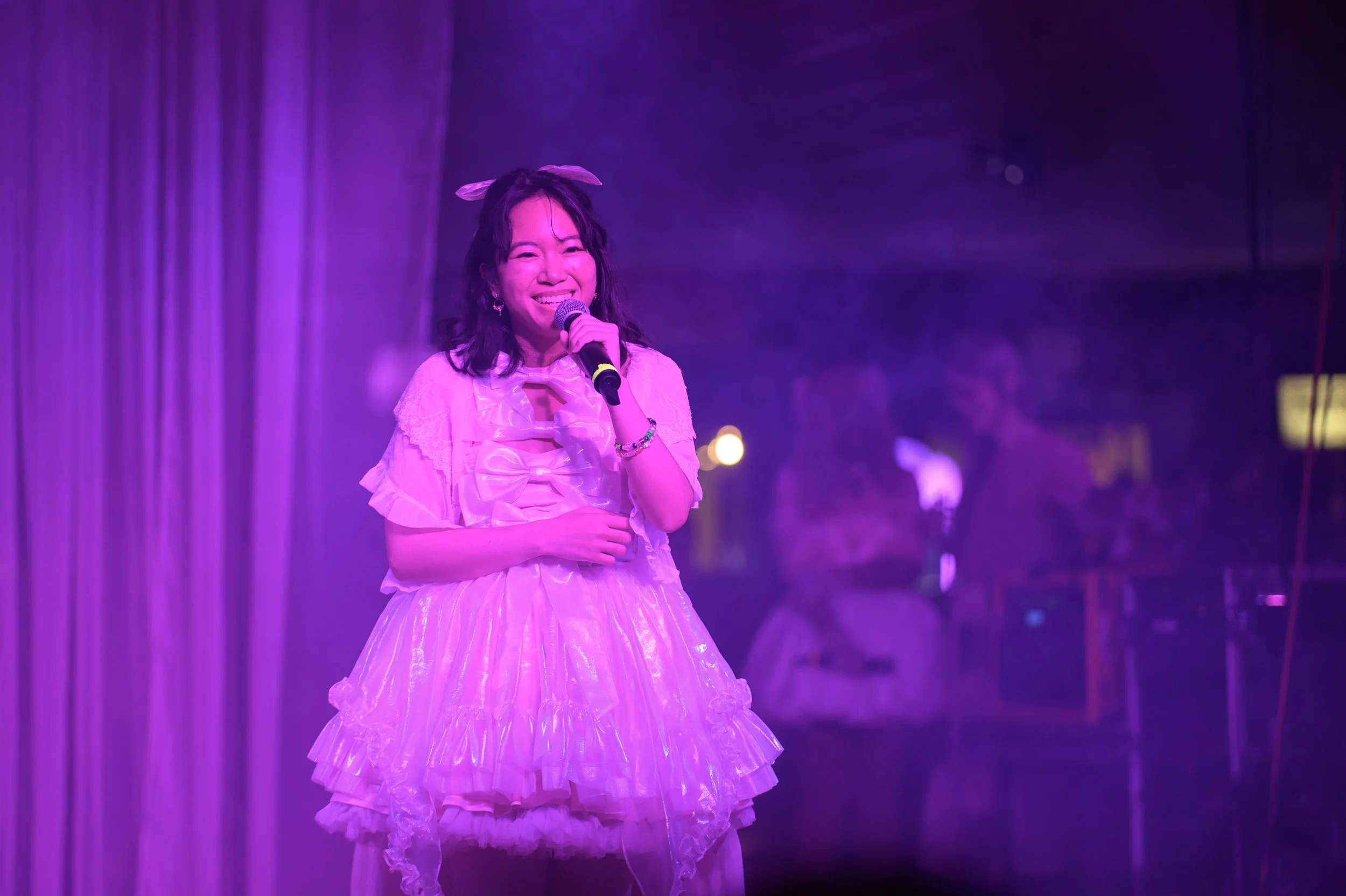A young woman in a frilly, pink dress with a bow detail, smiling and holding a microphone on stage with purple lighting and curtains in the background.