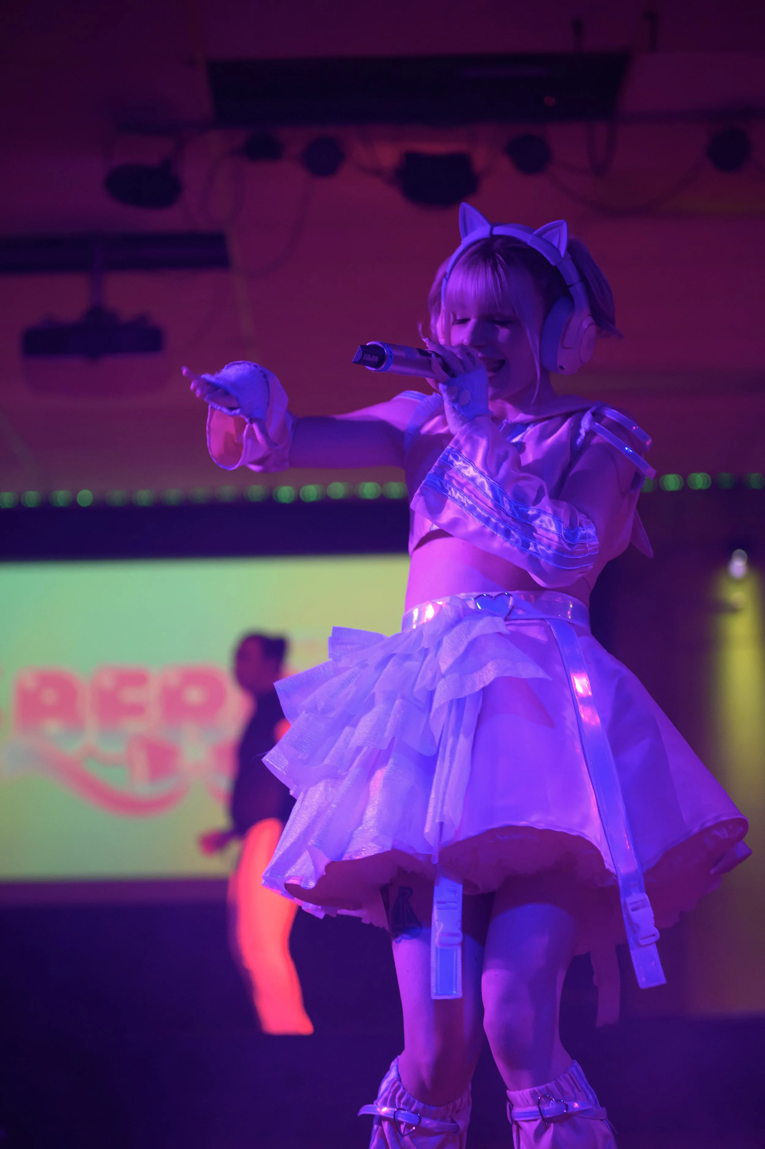 A performer dressed in a pink and purple costume with a tutu and cat ear headband, singing into a microphone on stage.