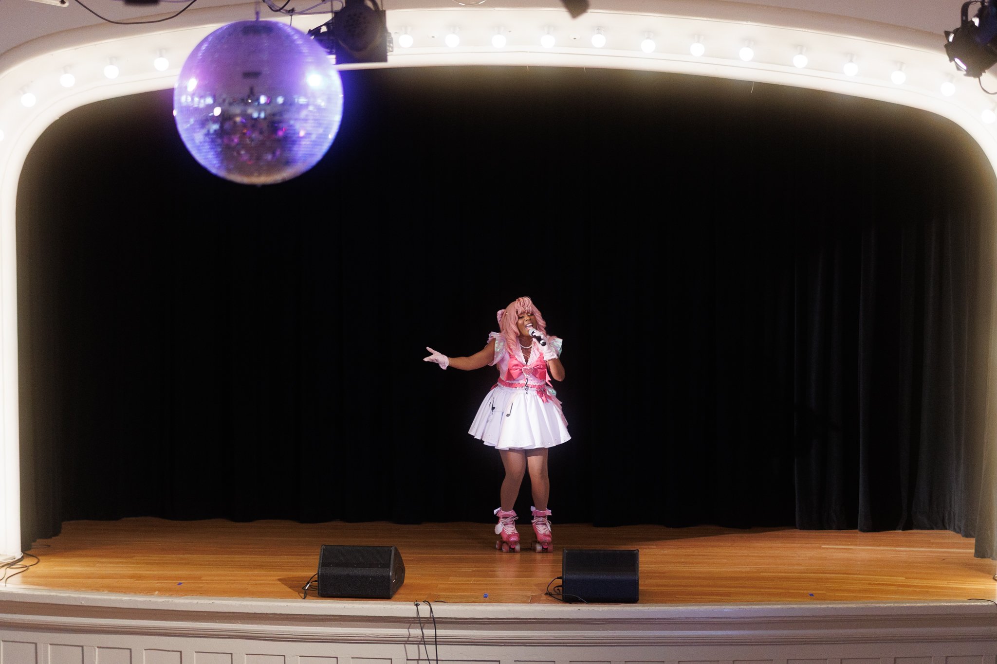 Performer singing on stage with pink hair, pink and white costume, and roller skates, under a disco ball with stage lights