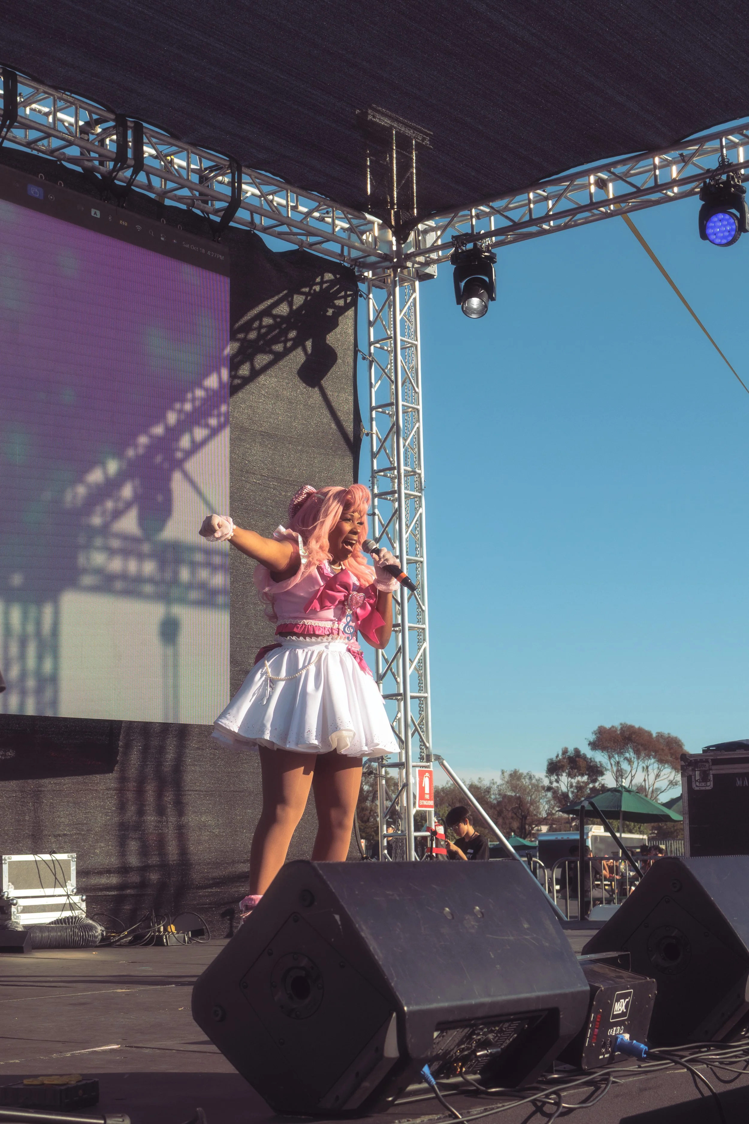 A performer with pink hair dressed in a pink and white costume singing on an outdoor stage under a clear blue sky.