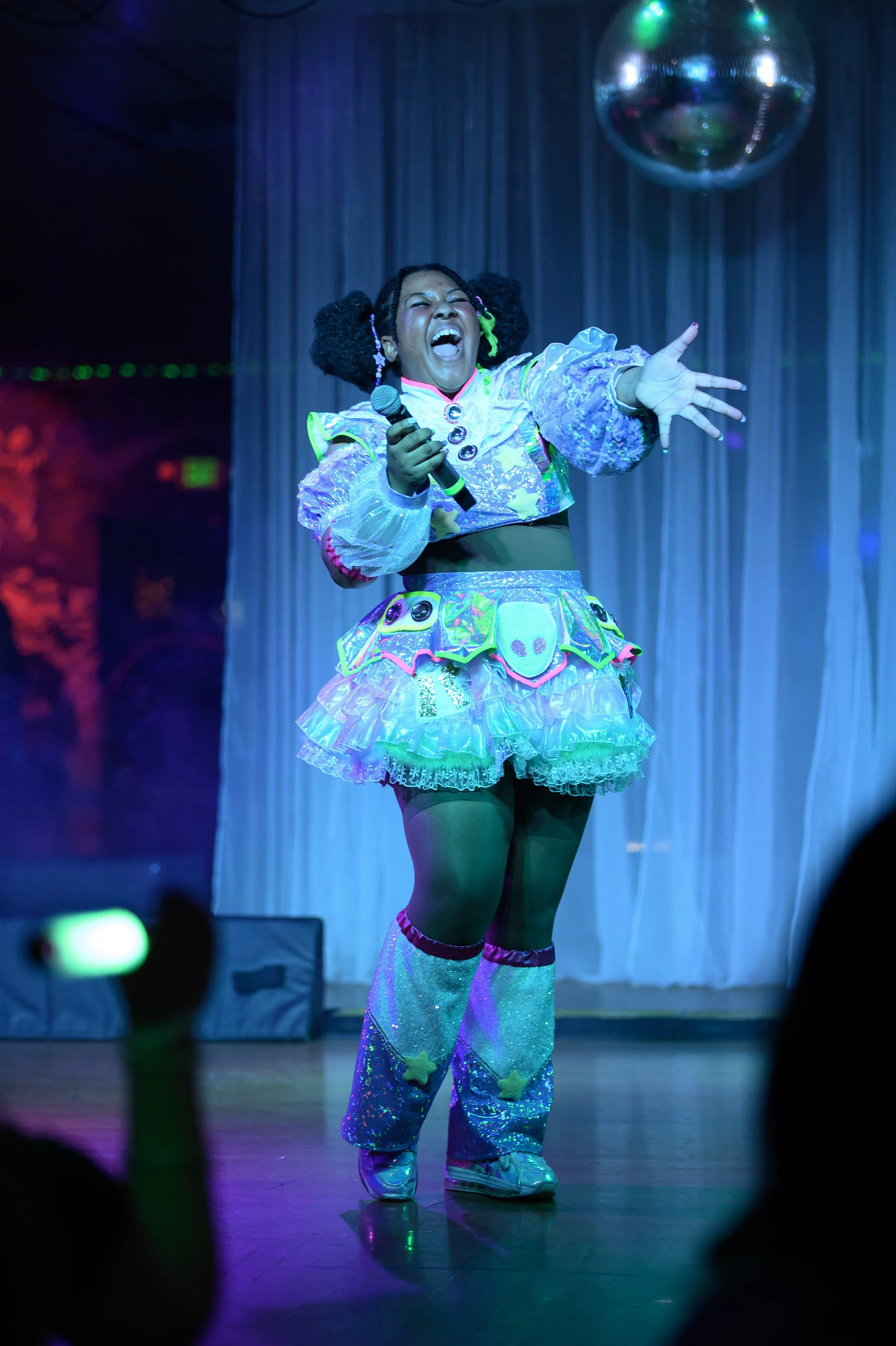 A performer singing or speaking on stage, wearing a colorful, holographic, and star-themed outfit, with a microphone in one hand, in front of curtains and a disco ball.