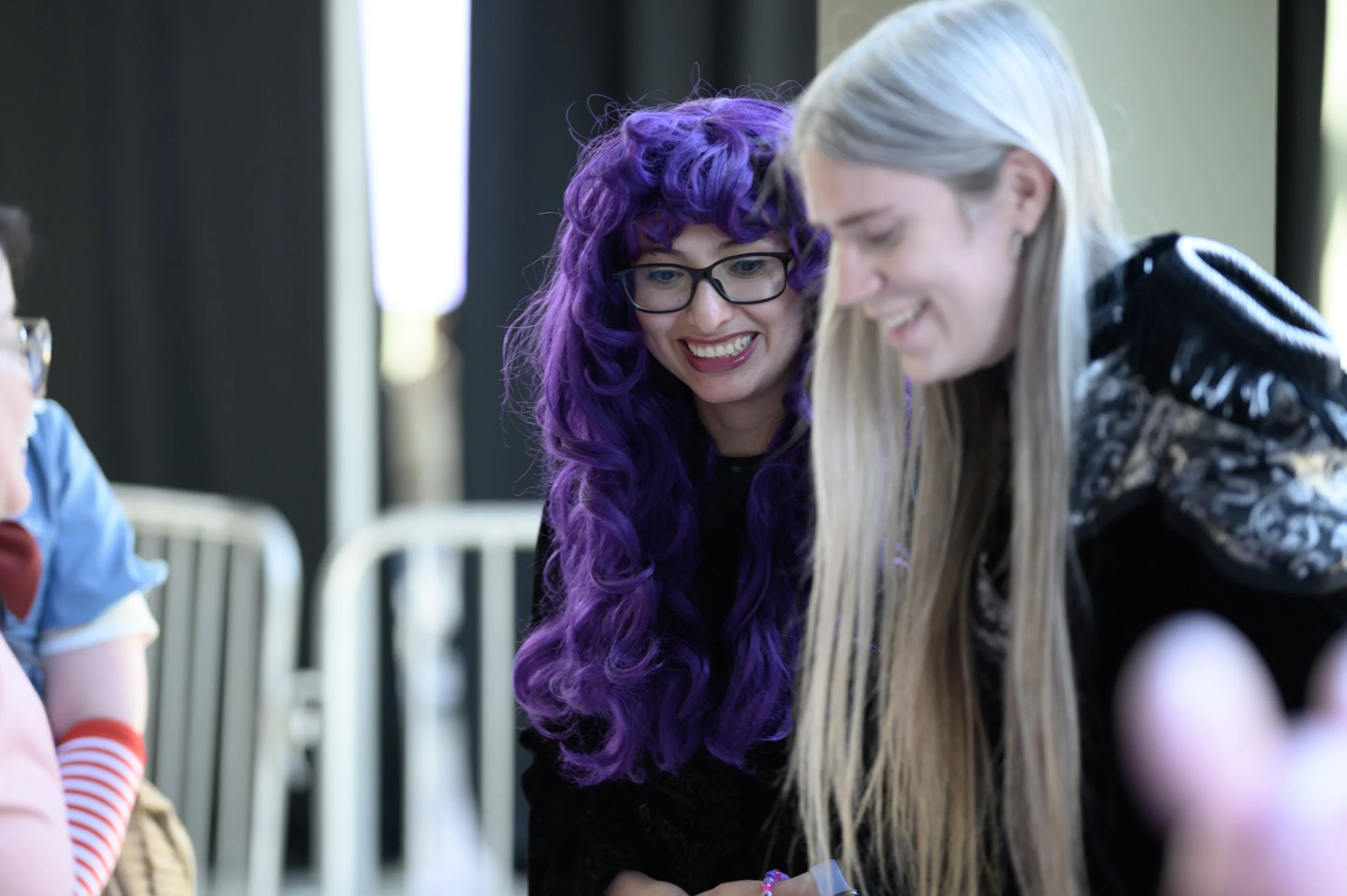 Two women smiling and looking at something off-camera indoors, one with long purple curly hair and glasses, and the other with long straight silver hair wearing a black jacket.