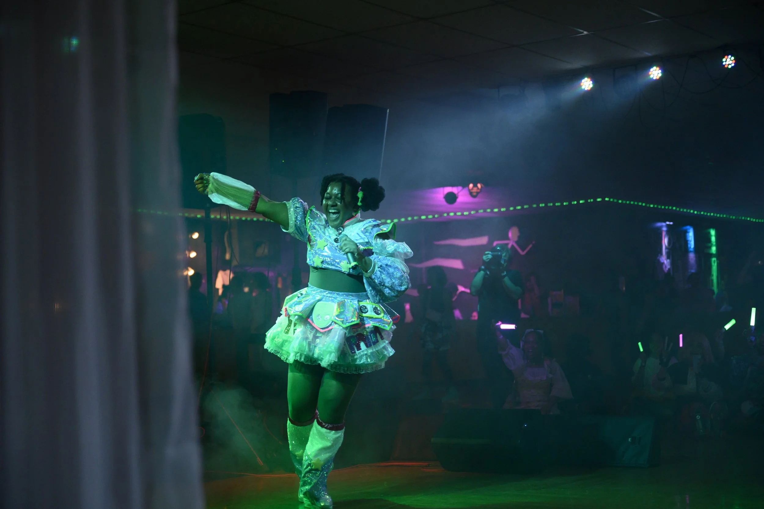 A performer on stage, wearing a colorful, shiny costume with a skirt and tall boots, holding a microphone and dancing in front of a dark audience with glow sticks, under vibrant stage lighting.