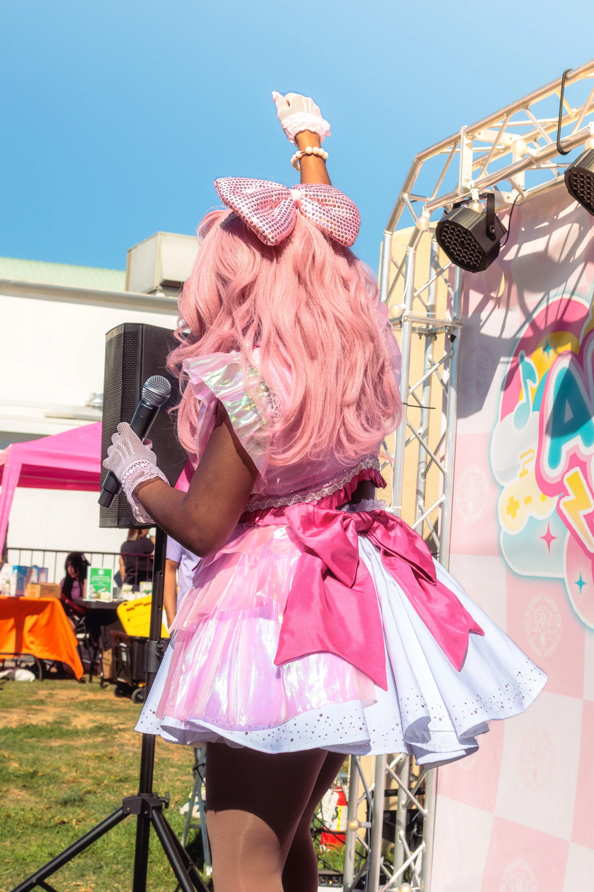 A woman dressed in a pink, shiny, frilly costume holding a microphone, with pink hair and a large pink bow in her hair, standing in front of a colorful stage at an outdoor event.
