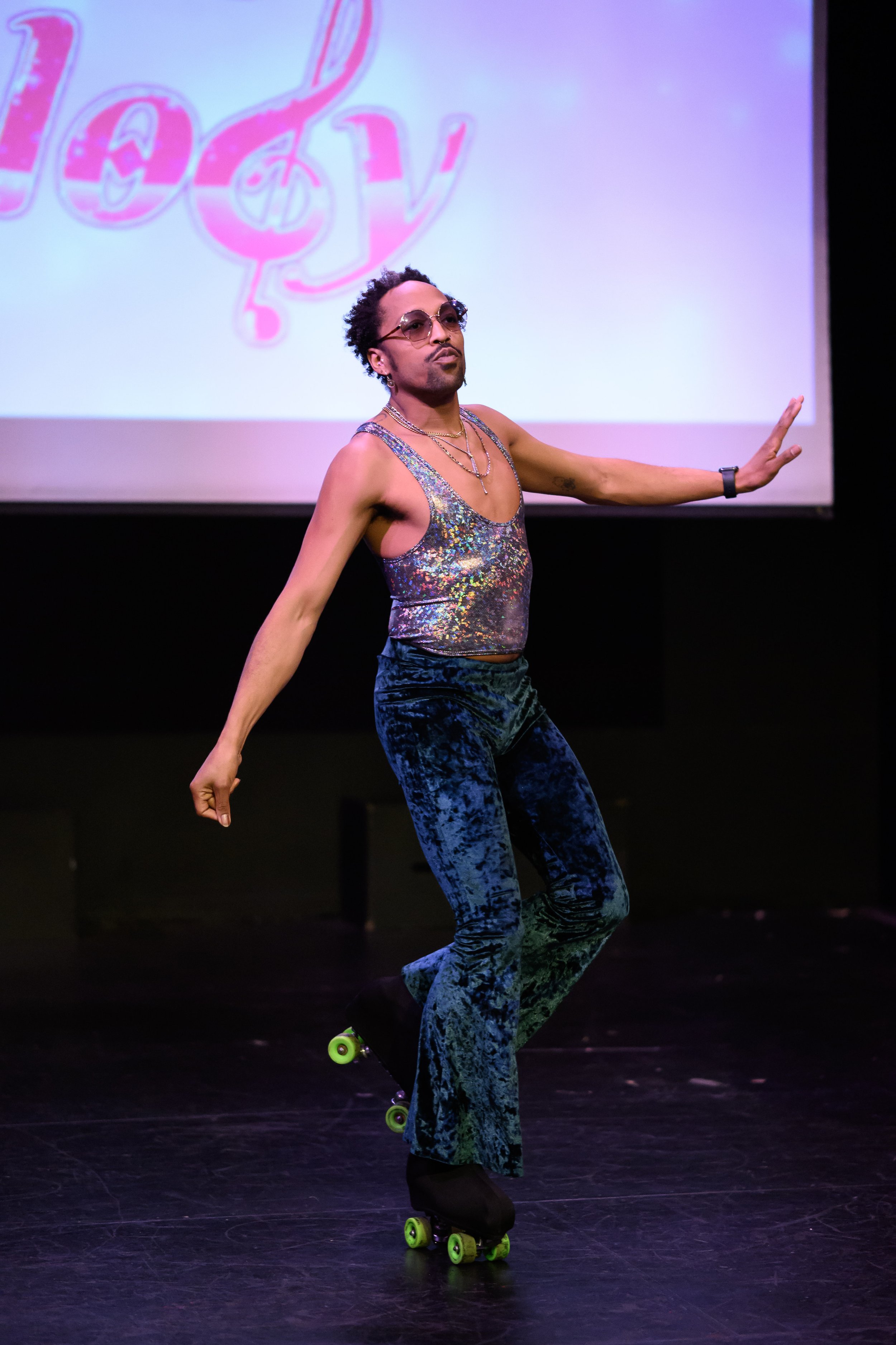 A person performing a dance or pose while on roller skates on stage, wearing a shiny, colorful tank top, blue velvet pants, sunglasses, and jewelry, with a large screen in the background displaying the word 'Melody'.
