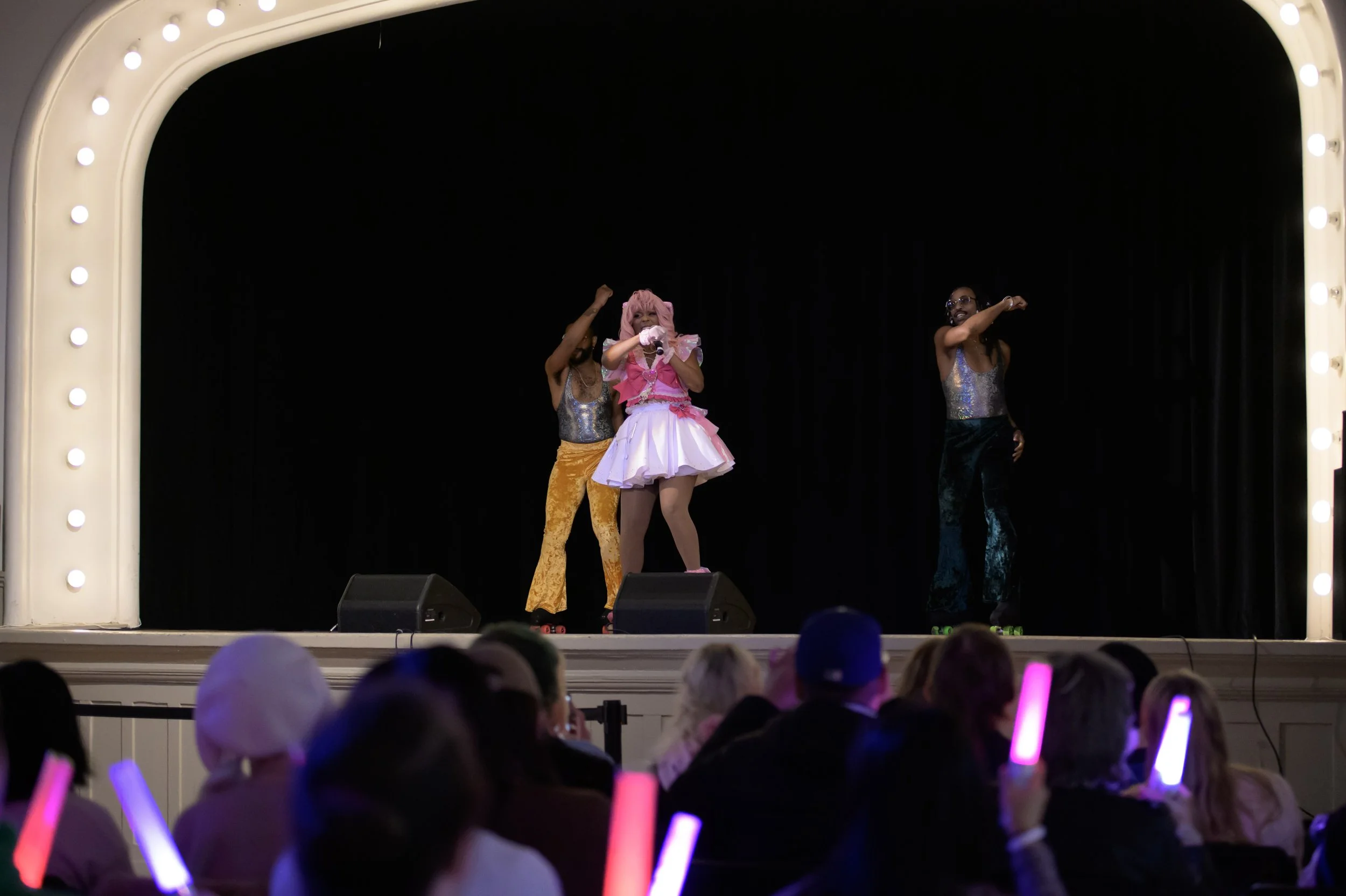 A stage performance featuring three performers, one dressed as a pink and white princess with a pink wig and others in shiny, colorful outfits, with an audience in front holding light sticks.