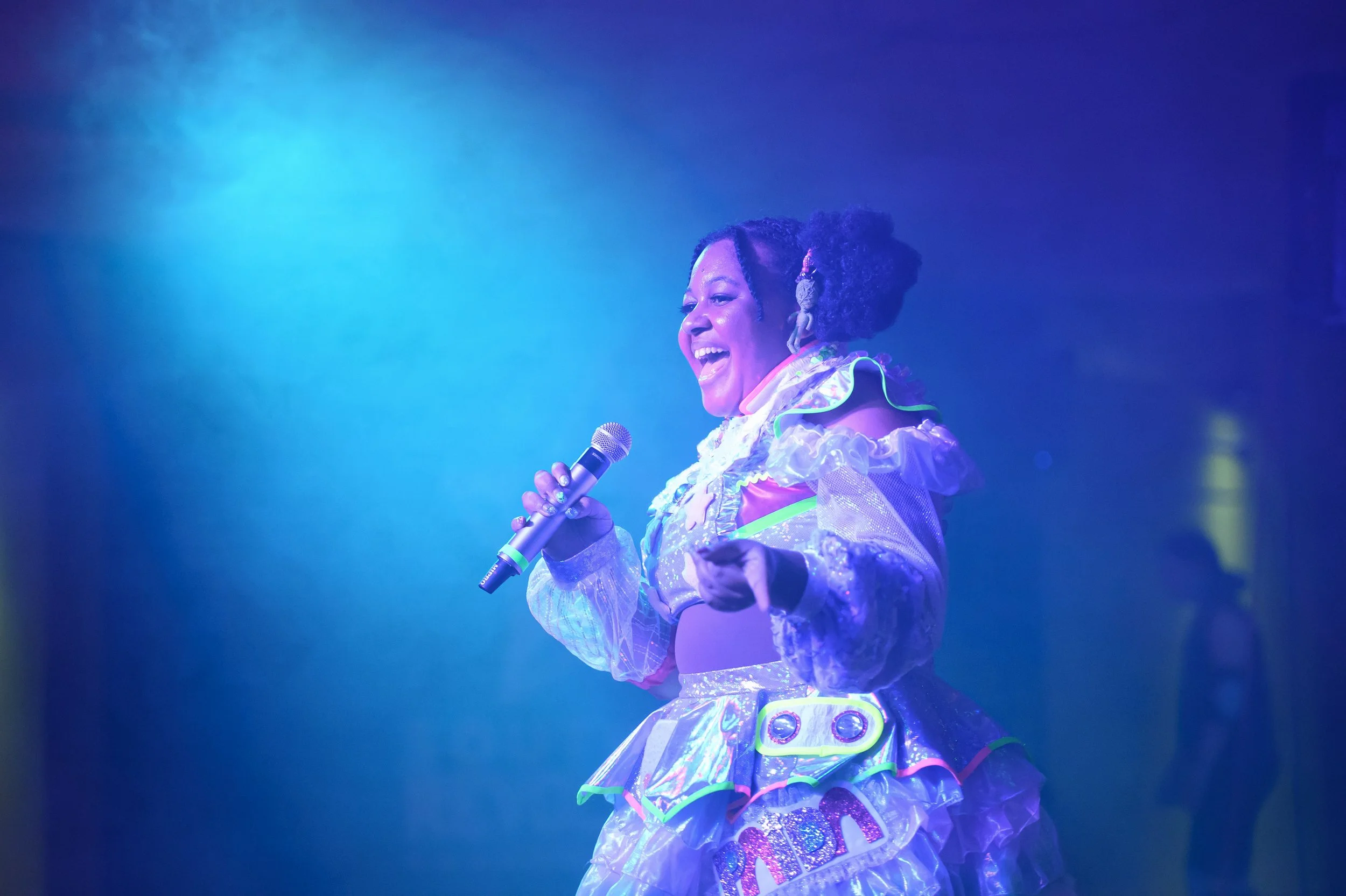 A woman on stage holding a microphone, wearing a colorful, holographic, futuristic costume with ruffled sleeves and a layered skirt, smiling with blue stage lighting in the background.