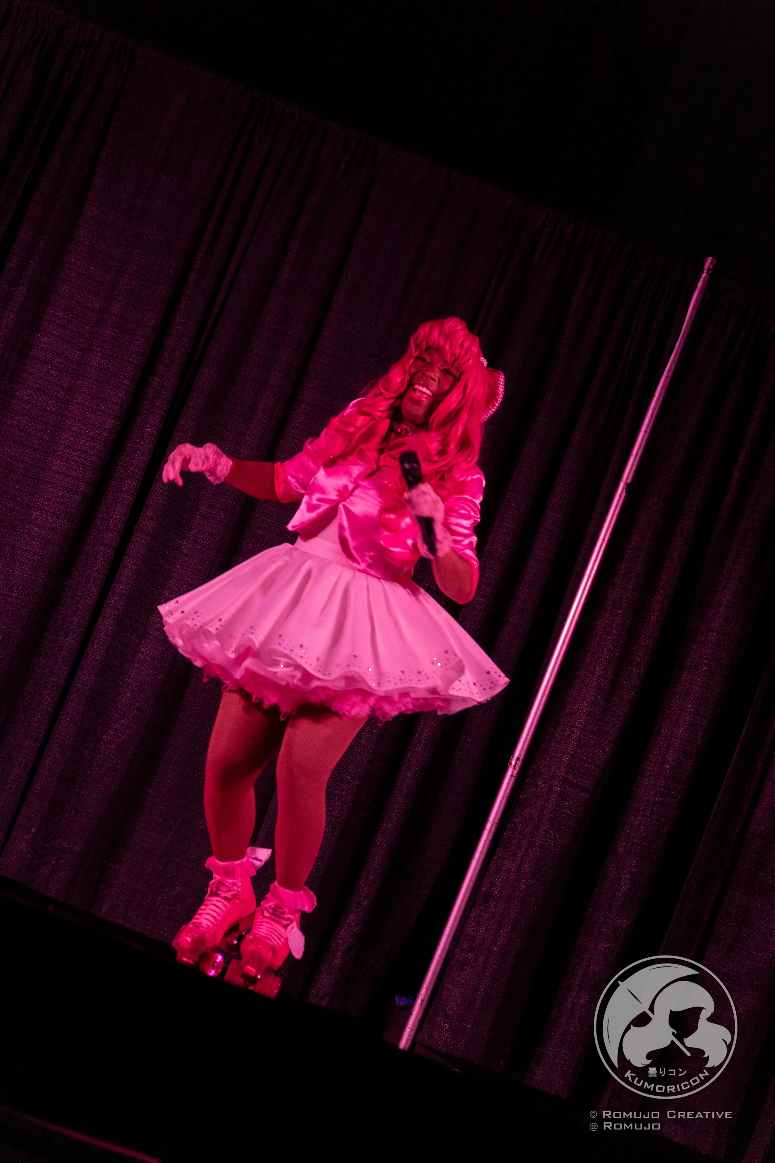 Performer dressed in pink with roller skates, holding a microphone, on stage with black curtain backdrop.