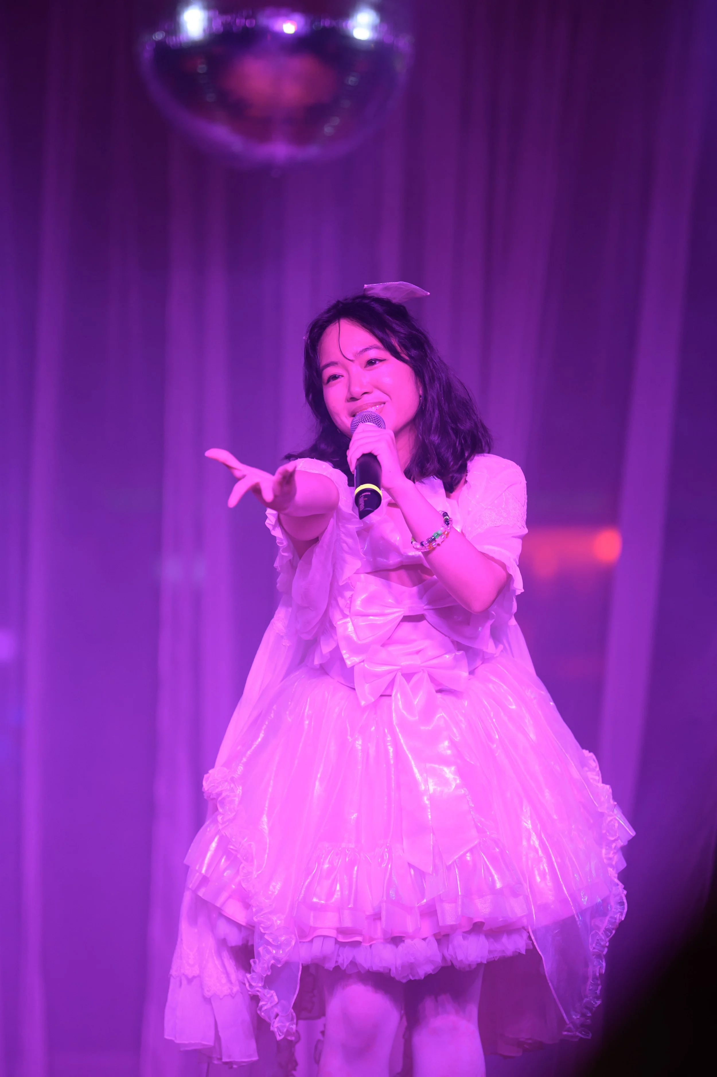 A young woman in a pink frilly dress singing on stage with a microphone, reaching out towards the audience, illuminated by purple stage lighting.