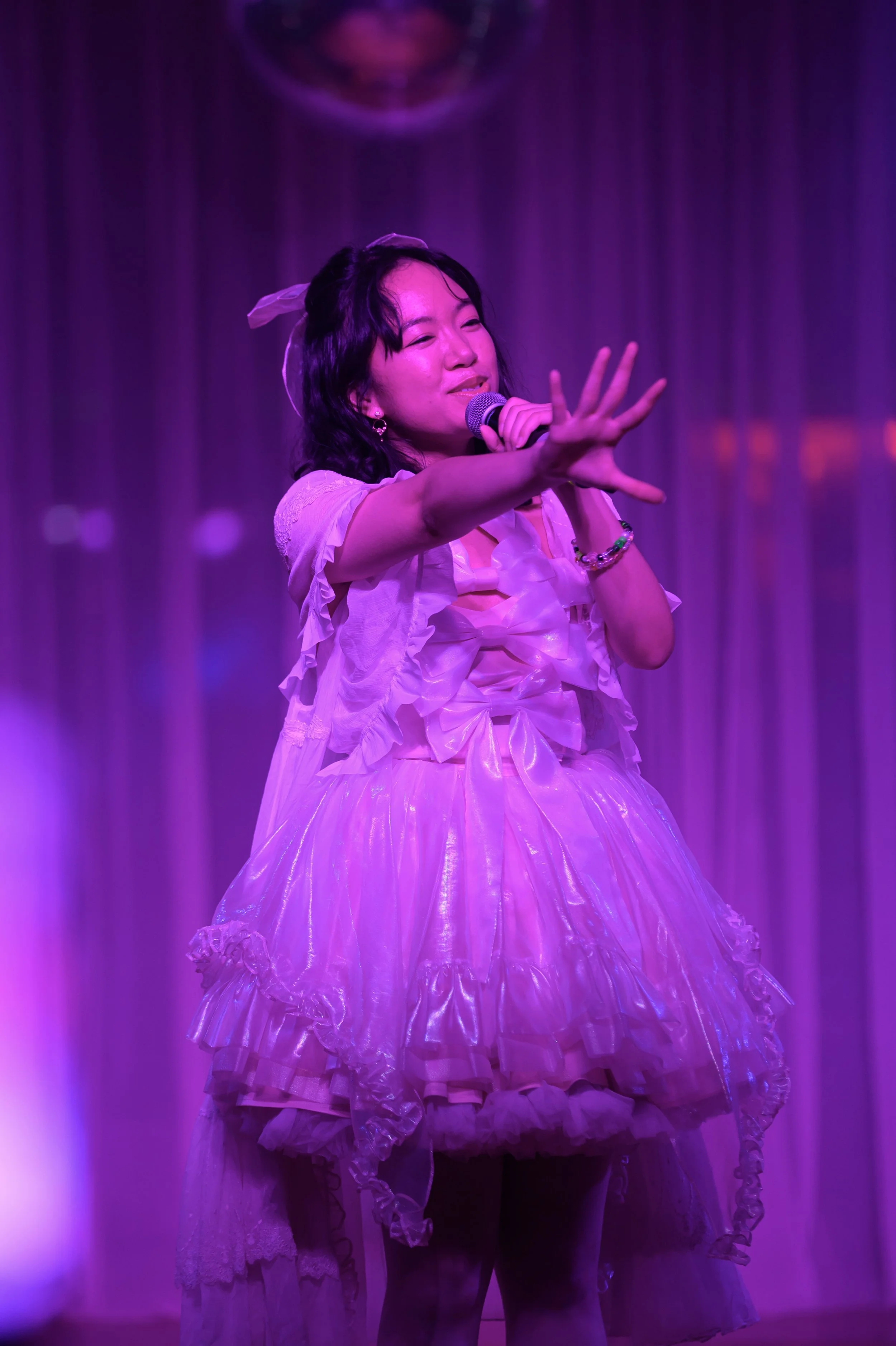 A young woman performing on stage, holding a microphone, dressed in a pink, ruffled, shiny dress with a bow, with purple lighting and a curtain backdrop.