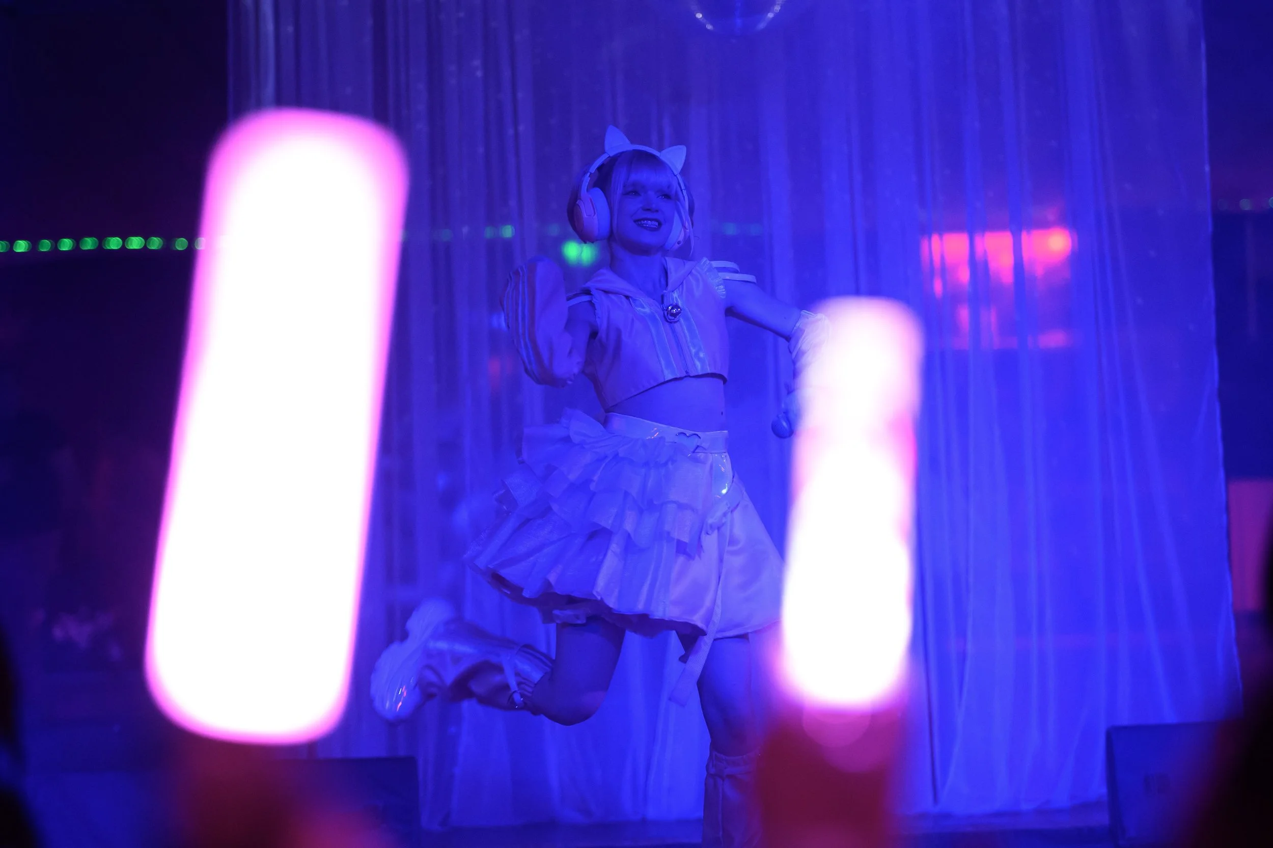 Person in a light-colored costume with cat ears, wearing headphones, dancing on stage with neon lights and curtains in the background.