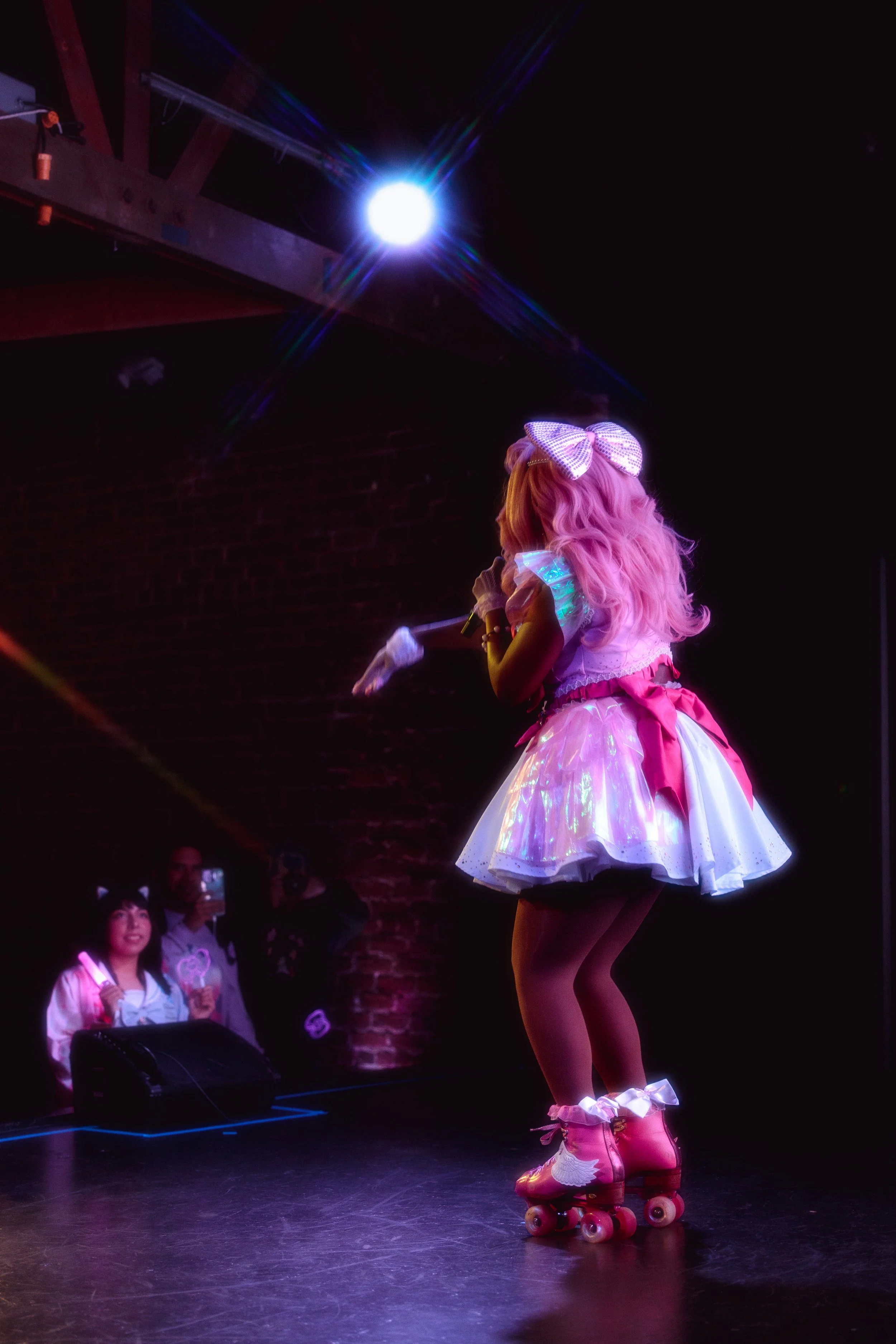 A girl in a pink and white dress with roller skates on stage, singing or speaking into a microphone during a performance, with two people watching in the background.