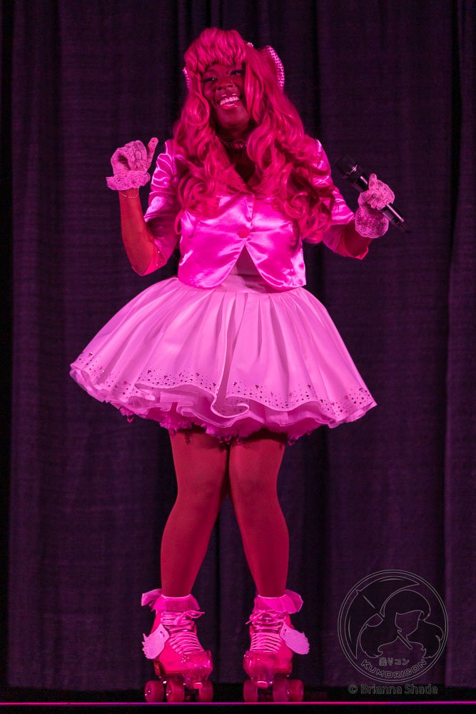 Person dressed in vibrant pink costume with roller skates, holding a microphone, standing on stage with dark curtain background.