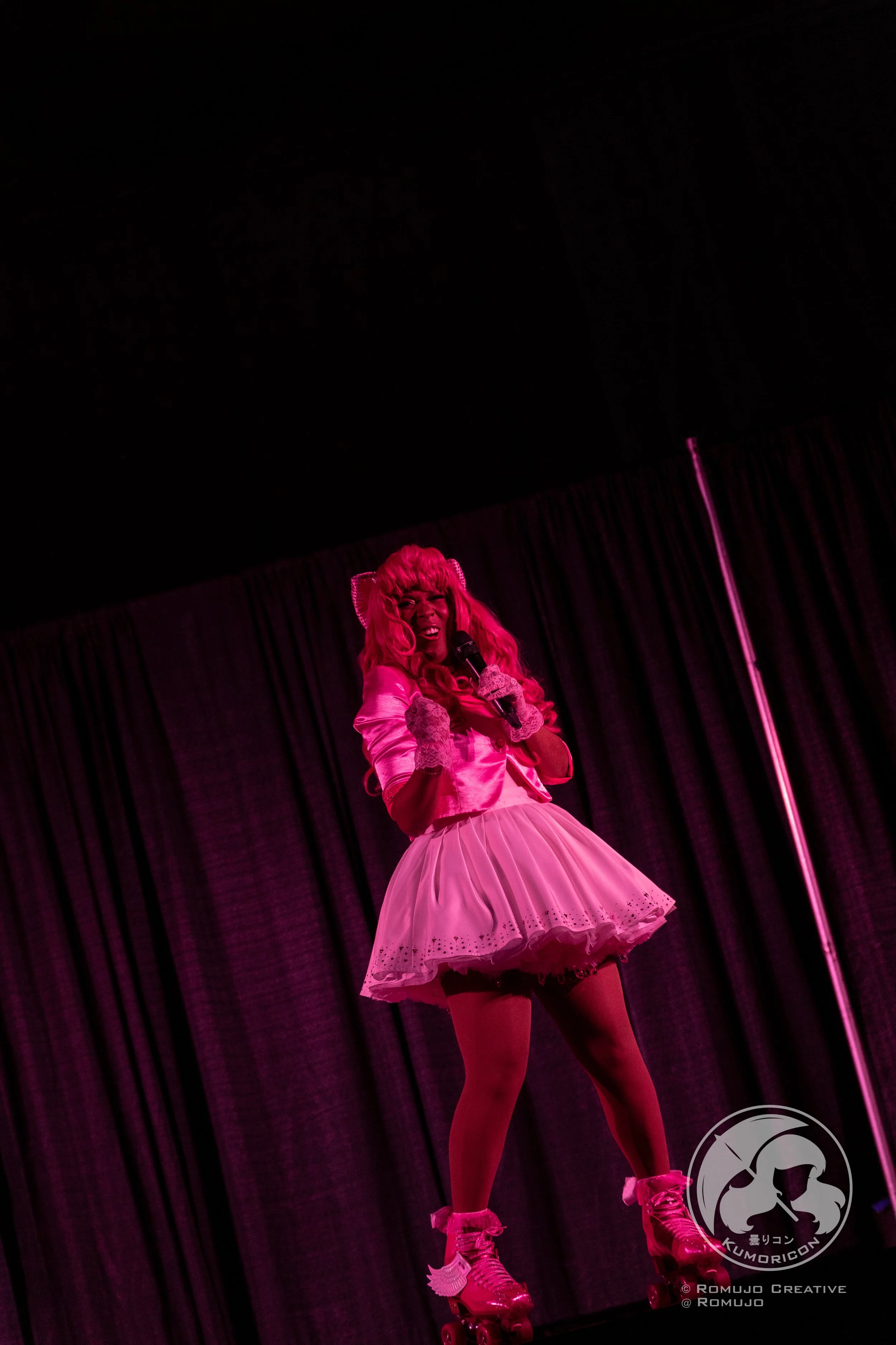 Performer dressed in pink with a wig, standing on stage, holding a microphone, in front of black curtains.