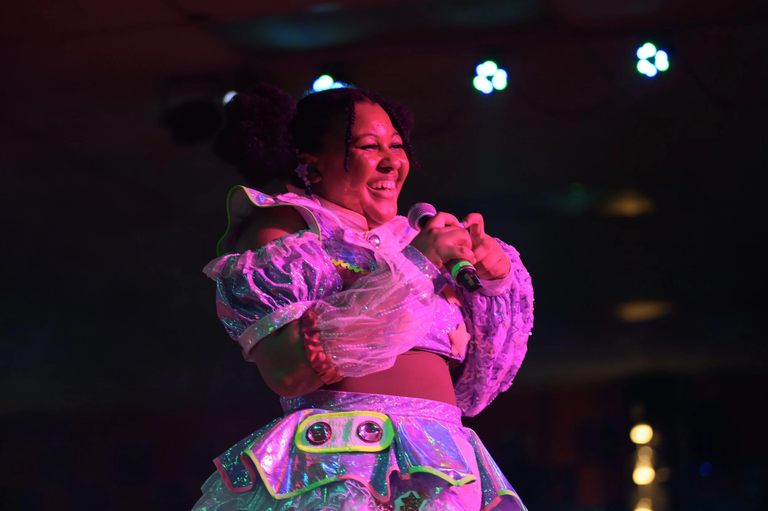 A woman with dark curly hair styled in puffs, smiling and holding a microphone on stage. She is dressed in a colorful, shiny, and ruffled costume with iridescent parts, illuminated by stage lights.