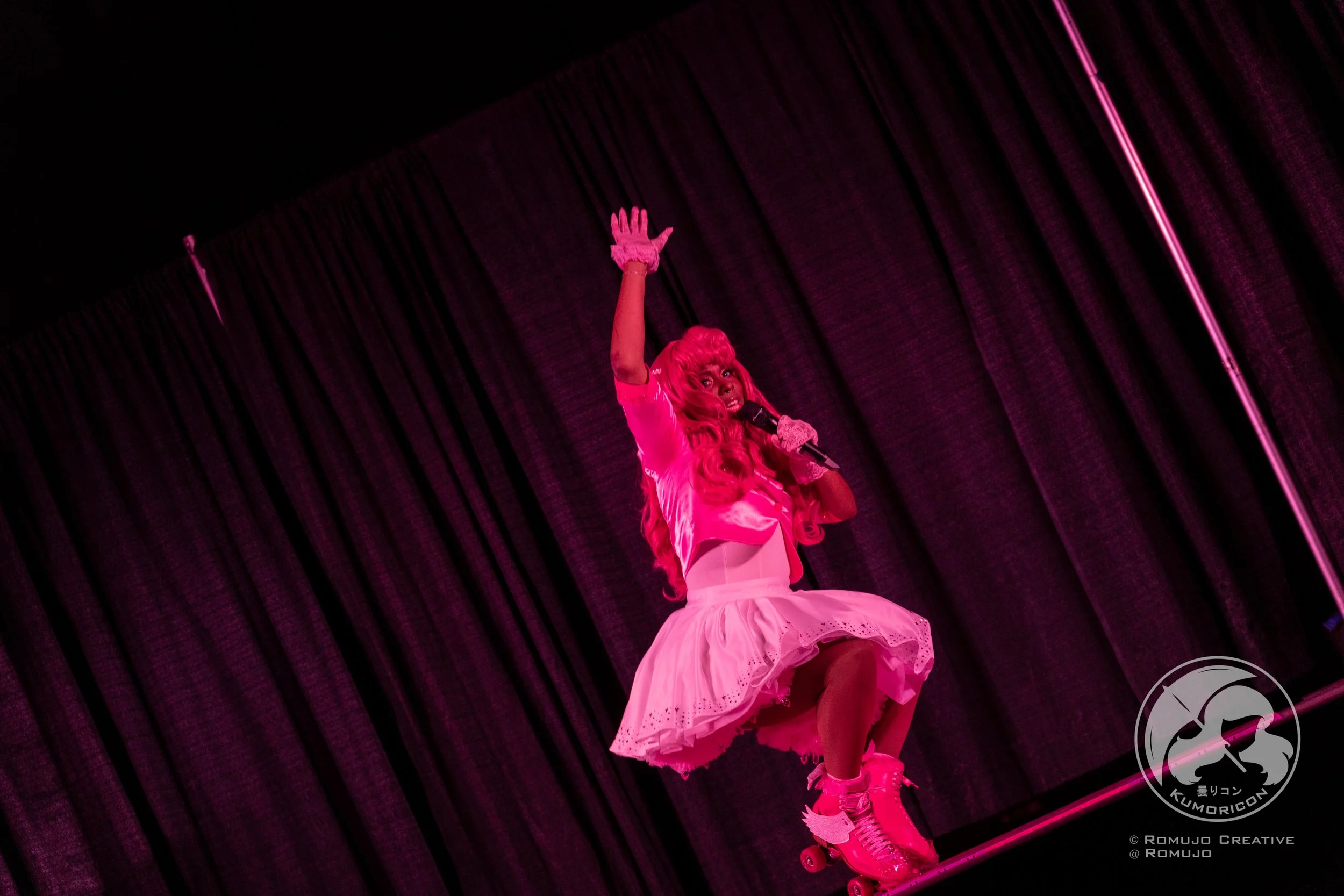 Performer dressed in pink and white, with a skirt and roller skates, on stage under pink lighting, holding a microphone and waving.