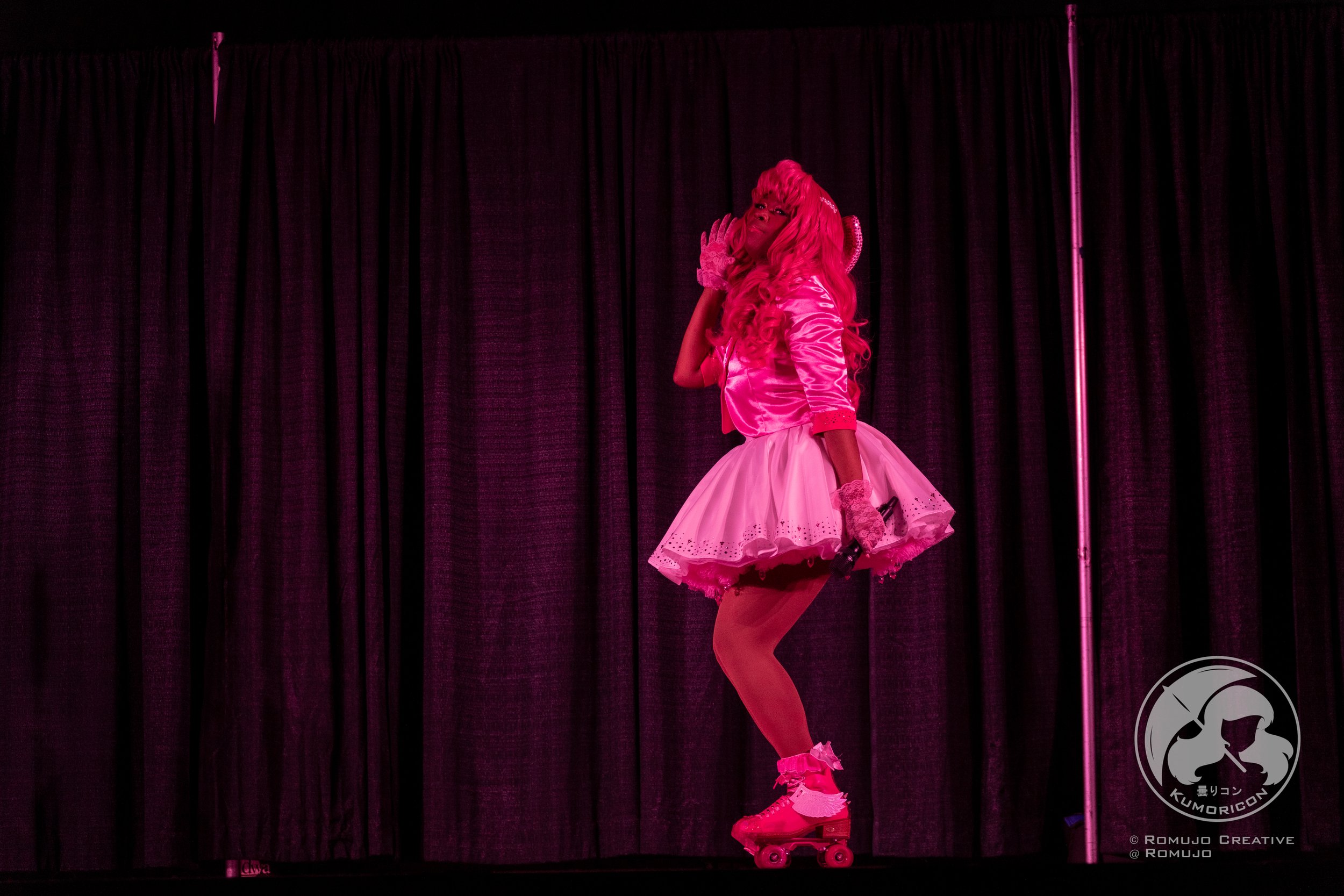 Person in pink wig, pink outfit, on roller skates, on stage with black curtains