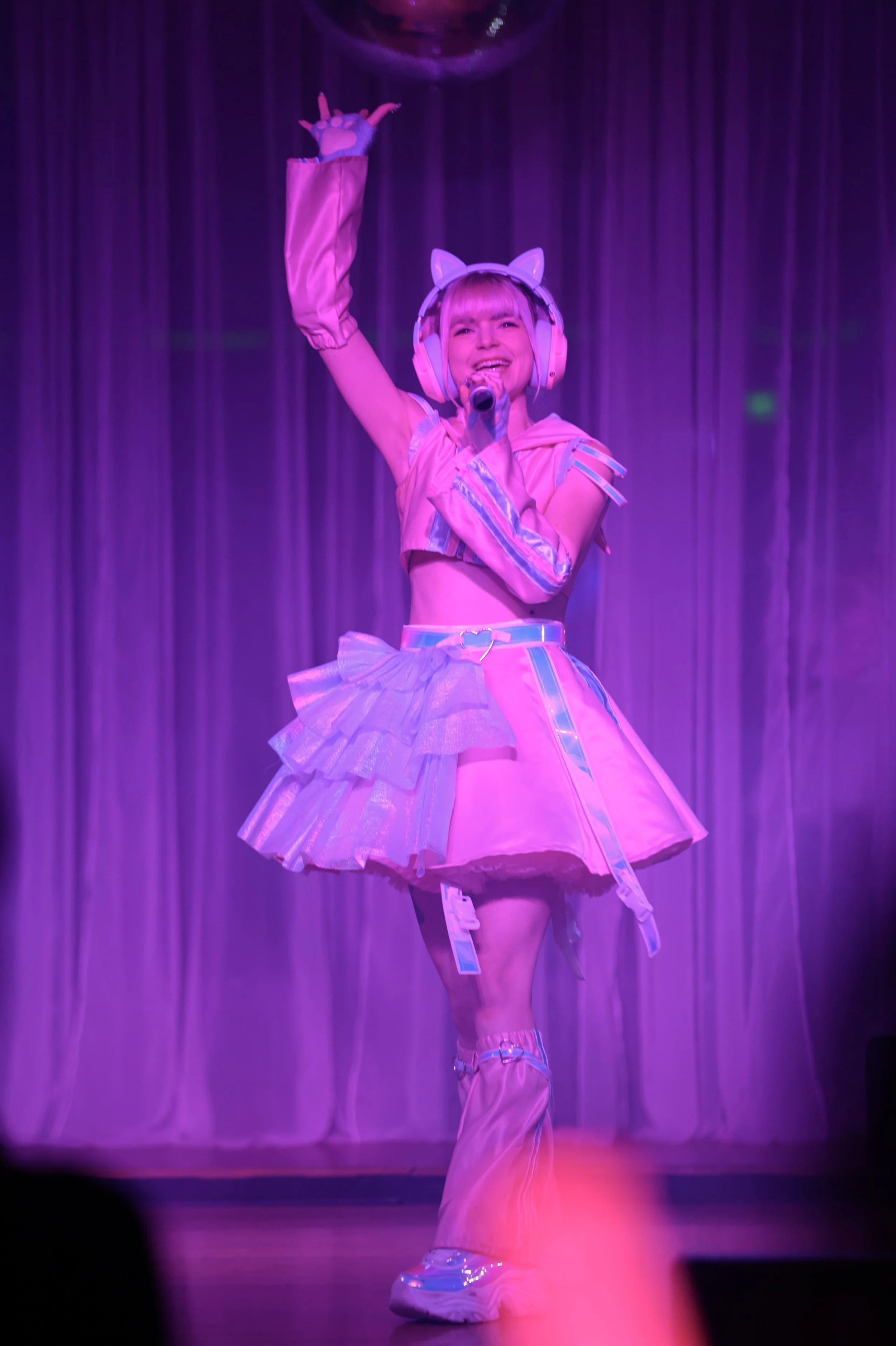 Performer dressed in a pink, futuristic, anime-inspired costume with cat-ear headphones, singing and dancing on stage under purple lighting.