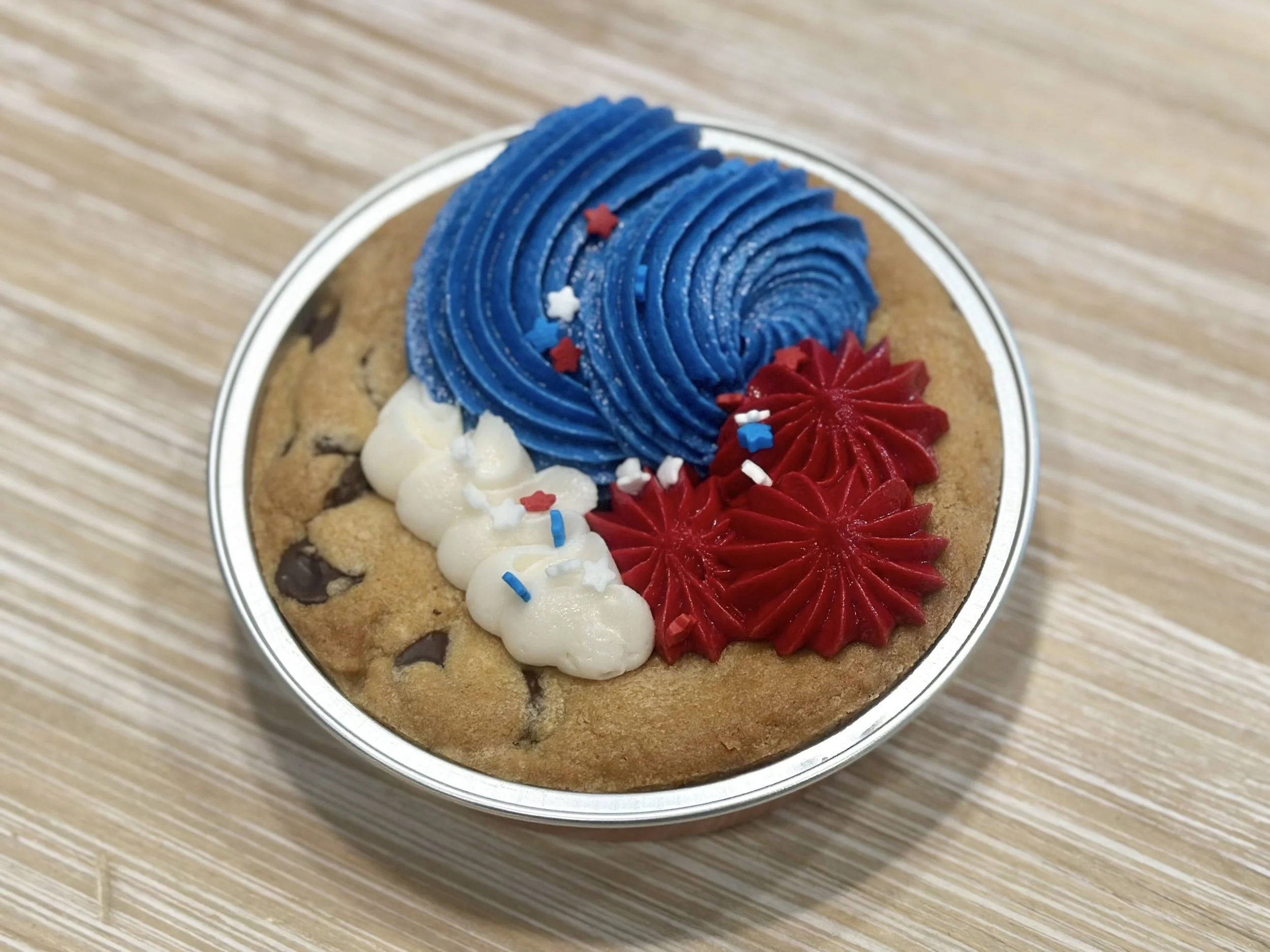 Chocolate chip cookie with red and blue whipped icing, white and red sprinkles, in a round metal container on a wooden surface.