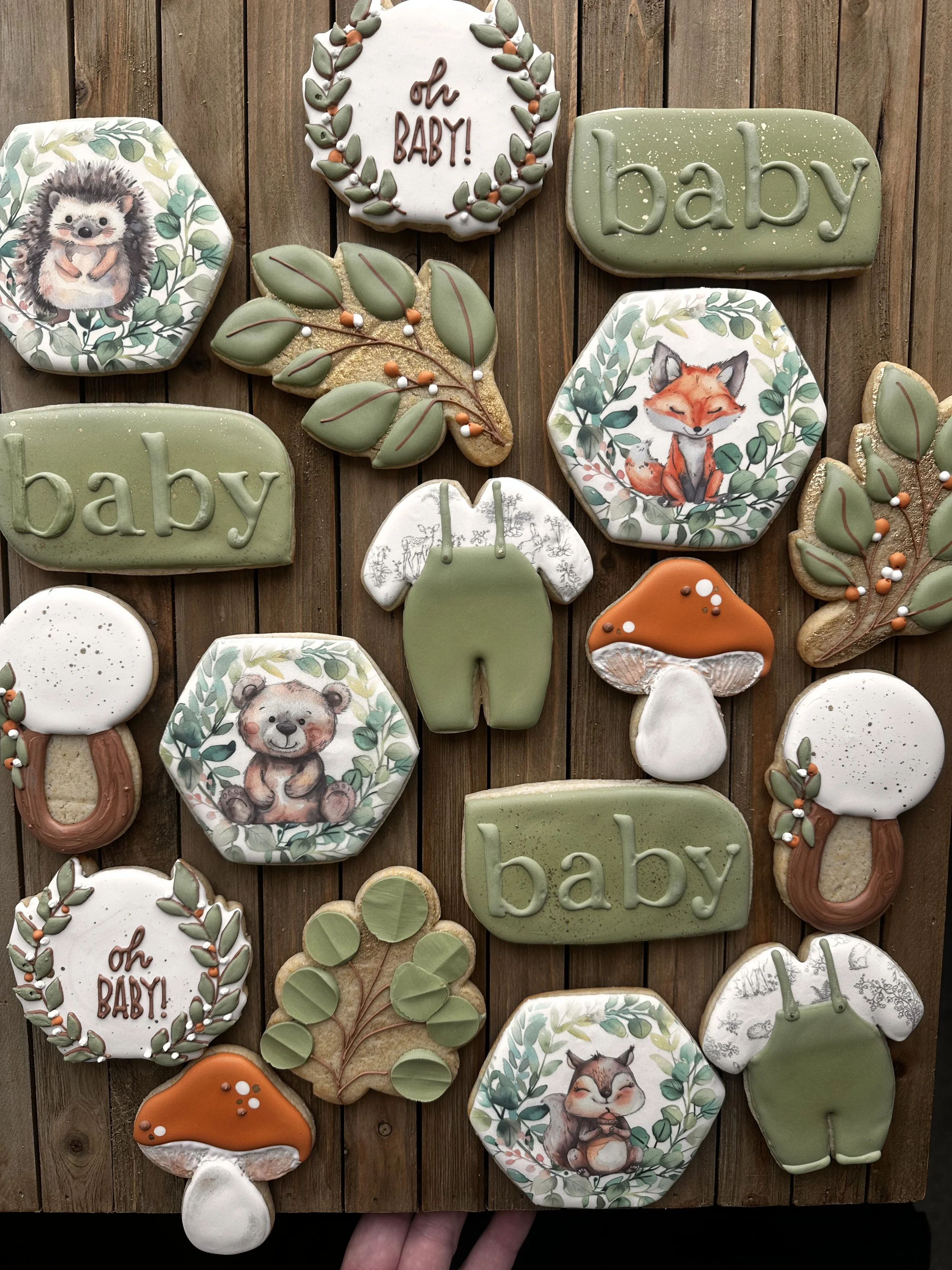 Assorted decorated cookies with baby, woodland animal, and nature themes, including hedgehogs, fox, bear, mushrooms, leaves, and signs saying "oh BABY!" and "baby" on a wooden surface.
