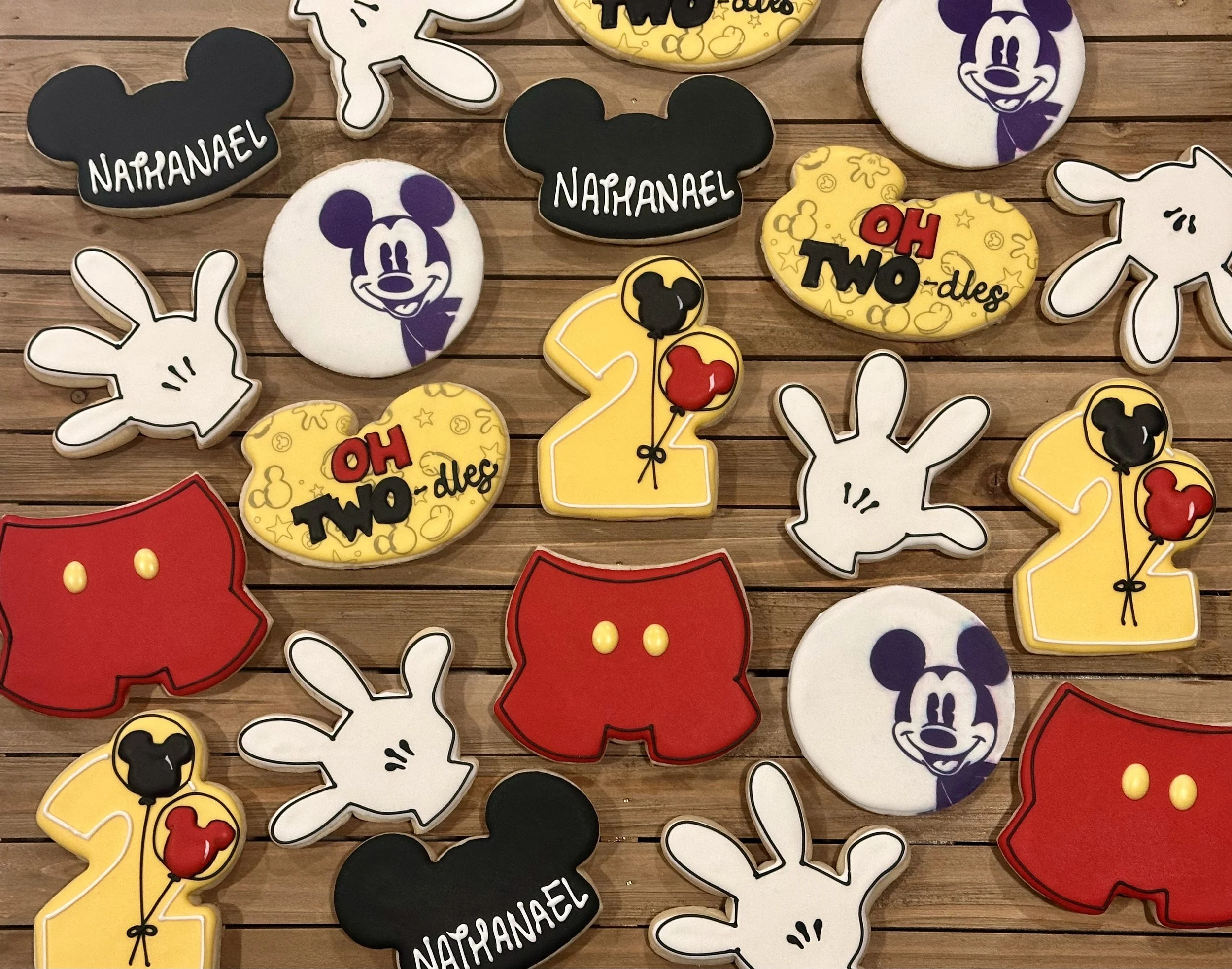 A collection of Mickey Mouse themed decorations and cookies on a wooden surface, including Mickey Mouse ears, white gloves, red shorts, balloons, and yellow number 2 with balloons, featuring cartoon and party design elements.