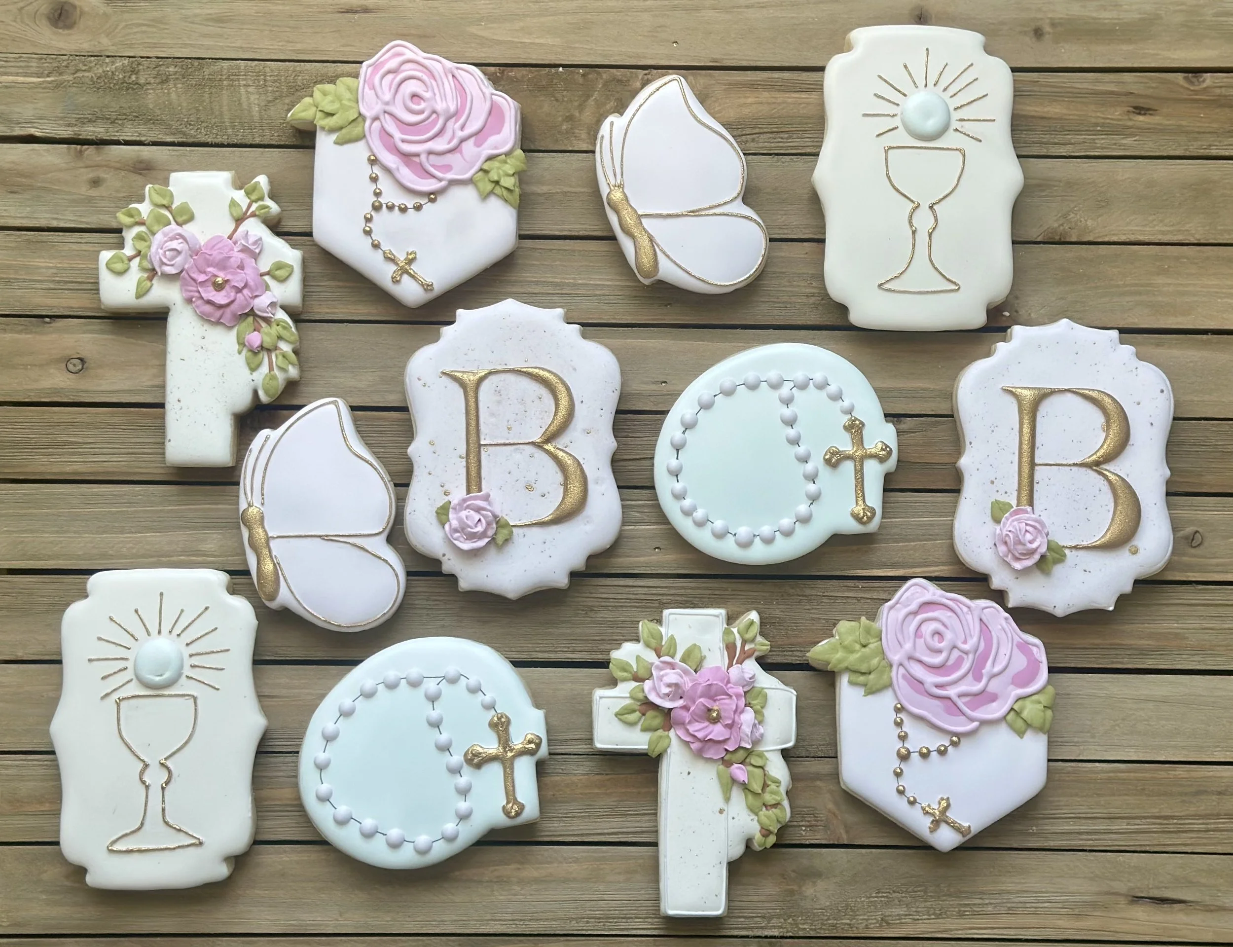 Assorted decorated religious cookies on a wooden surface, including crosses, roses, chalices, prayer beads, and dove motifs, with a white, pink, and gold color scheme.
