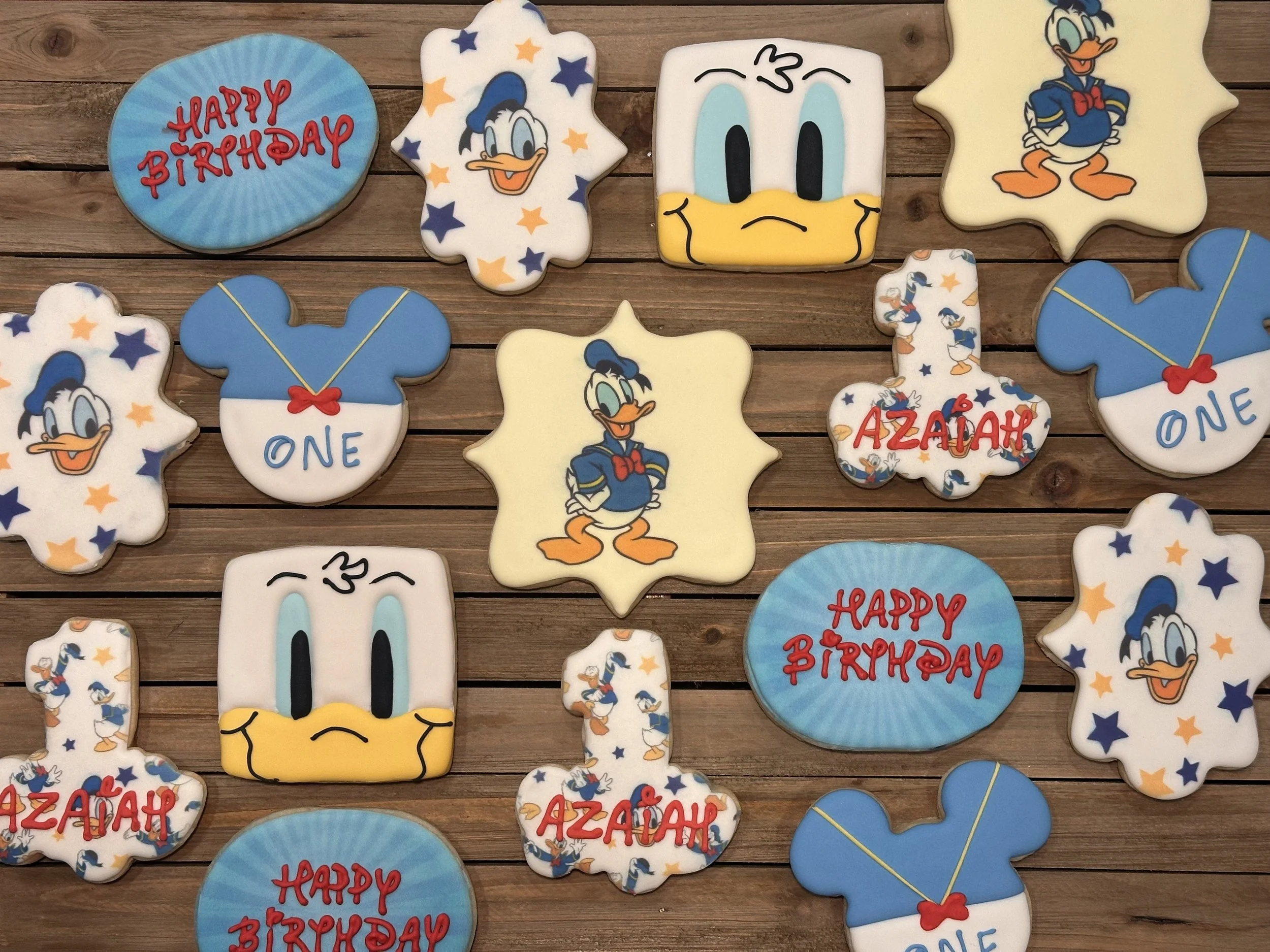 Decorative cookies with Donald Duck theme, including words 'Happy Birthday' and 'One', and images of Donald Duck and his signature hat, on a wooden surface.
