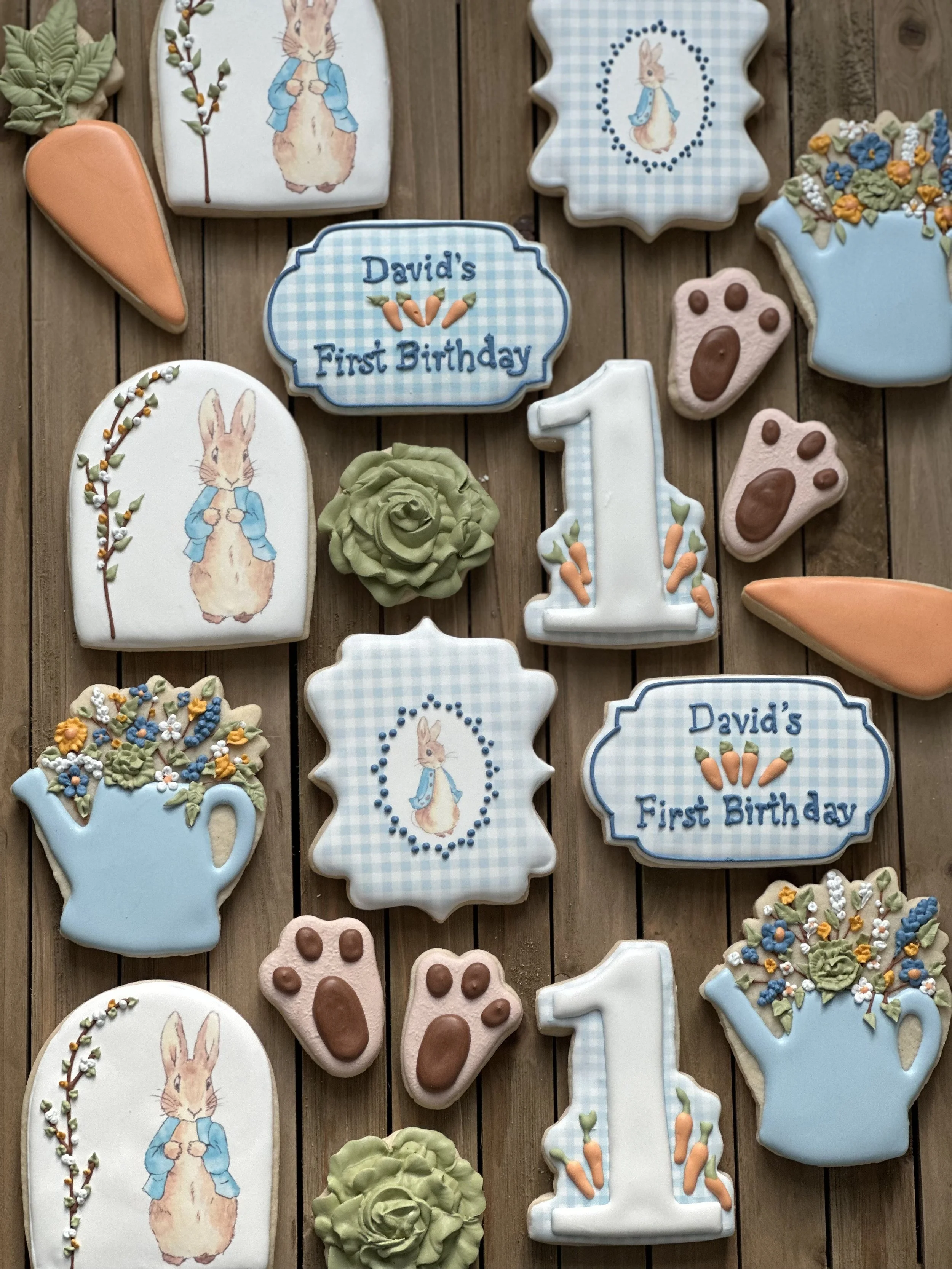 Assorted decorated cookies on a wooden surface: rabbits in blue jackets, carrot shapes, pink paw prints, blue watering cans with flowers, a green rose, a number one with carrots, and signs reading "David's First Birthday".