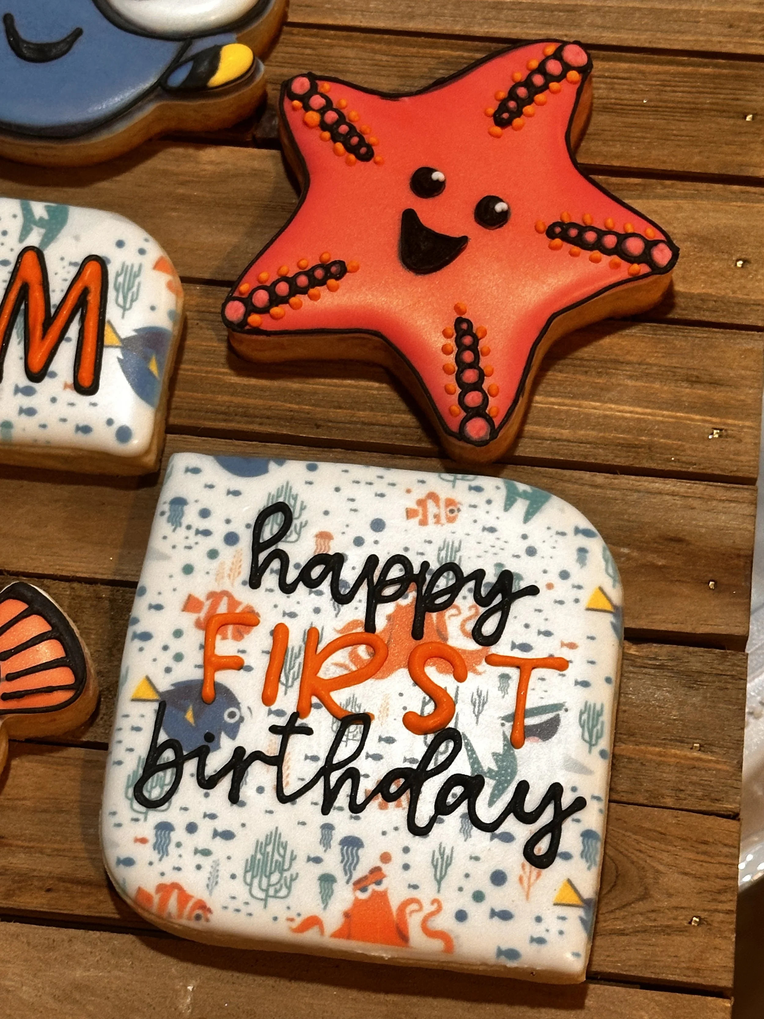 Decorated birthday cookies with a starfish face cookie on a wooden table and a square cookie that says "happy FKCST birthday" with ocean-themed design.