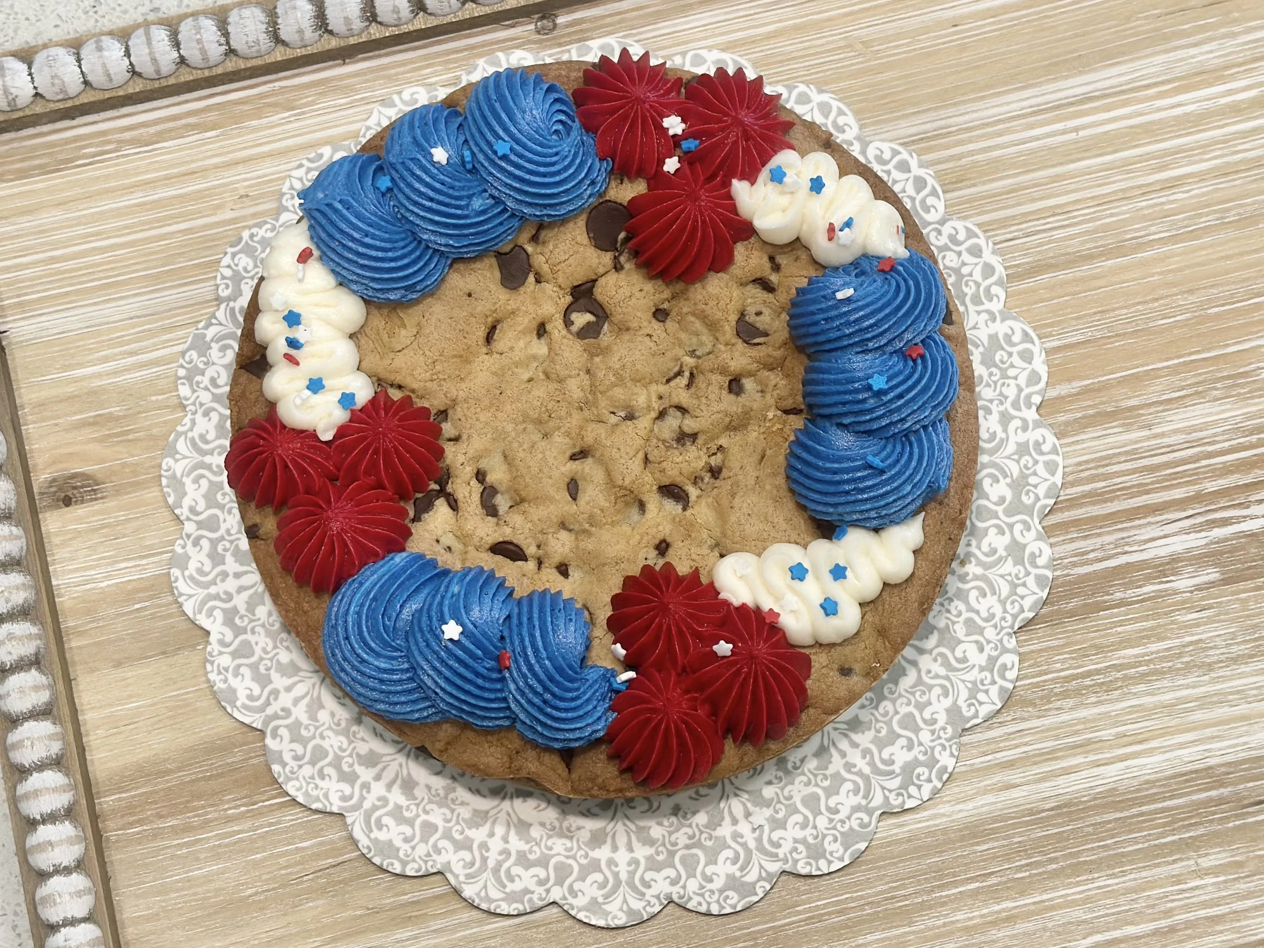 Decorated chocolate chip cookie with red, white, and blue frosting and star-shaped sprinkles on a decorative paper doily.