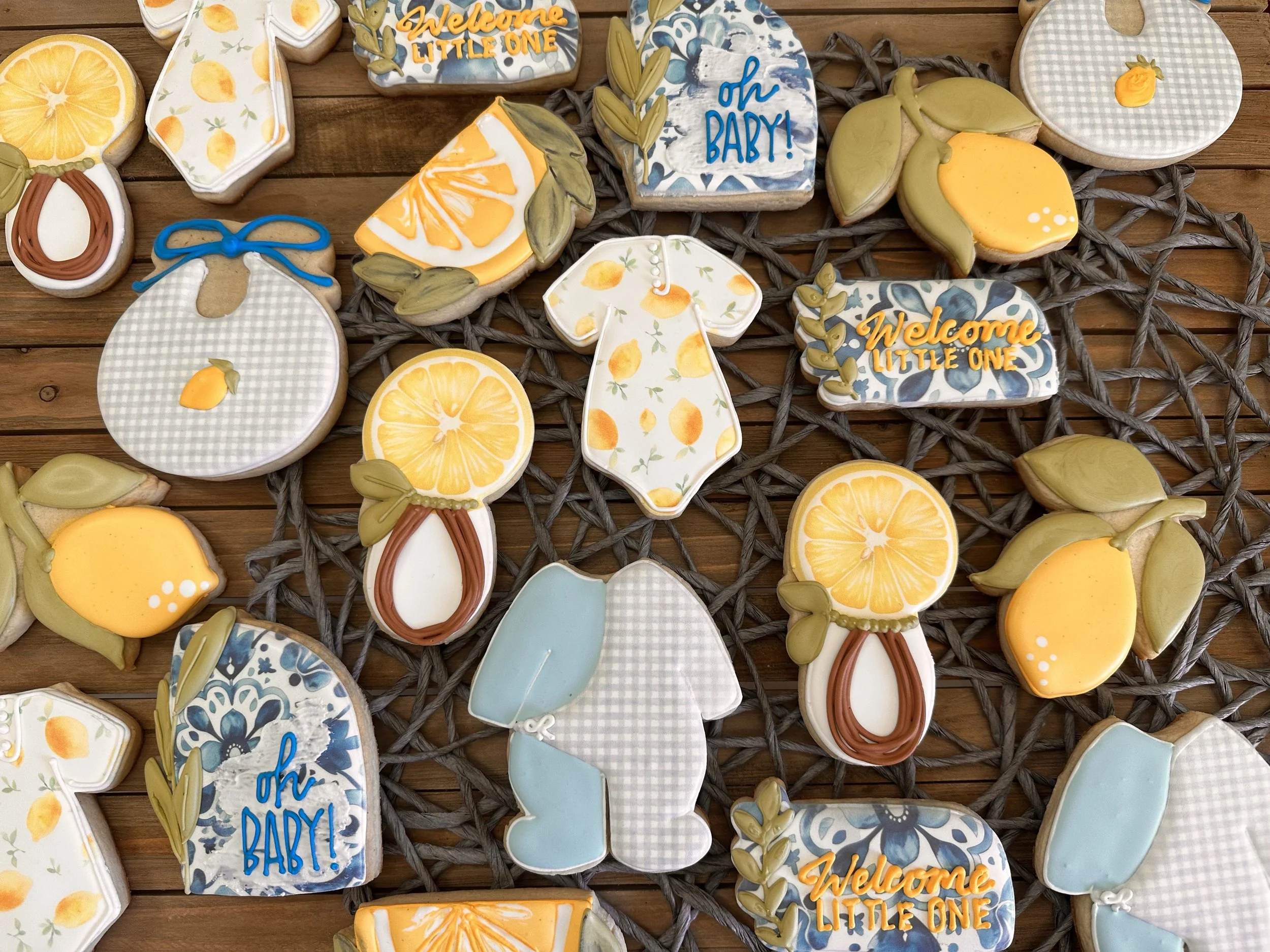 Collection of decorated cookies with lemon-themed and baby shower designs, featuring phrases like 'Welcome Little One' and 'Oh Baby,' on a wooden surface.