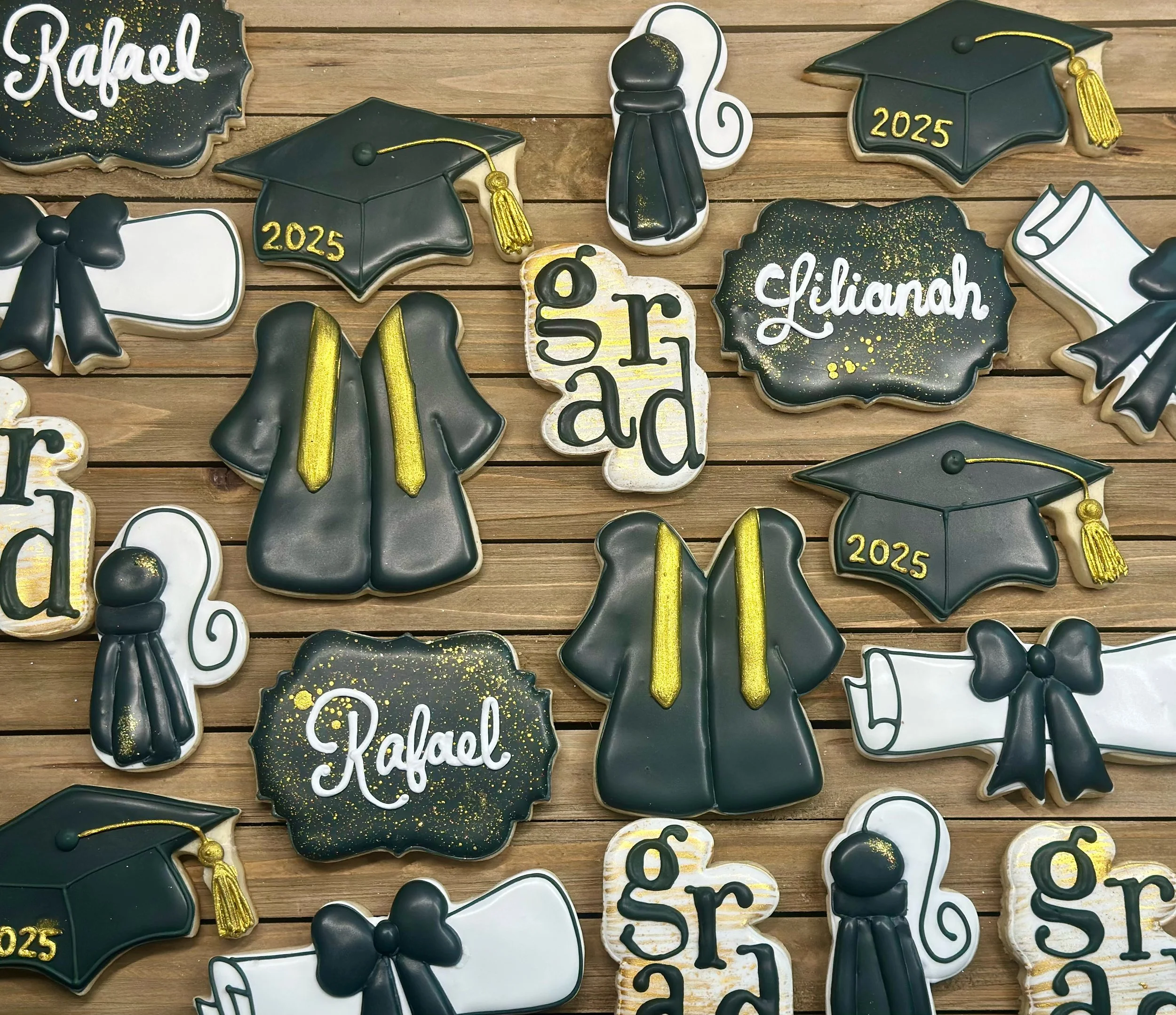 Decorated cookies for graduation, including graduation caps, diplomas, gowns, and the words 'graduate', 'Rafael', 'Liliana' in white script, with black, white, gold, and gray colors, arranged on a wooden surface.