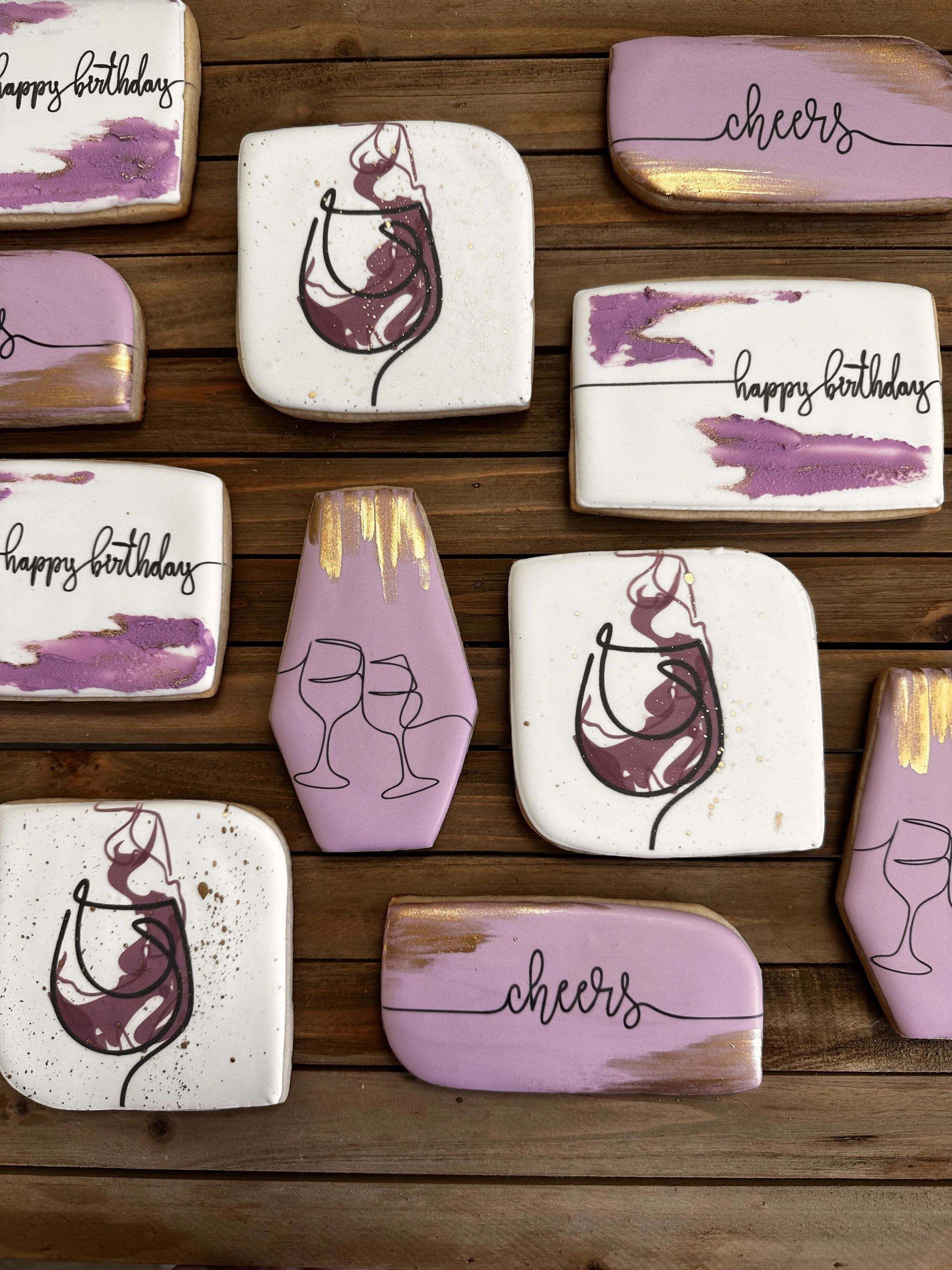 Decorated cookies on a wooden surface, some with the words 'happy birthday' and 'cheers,' others with illustrations of wine glasses and a wine glass outline with purple and gold accents.