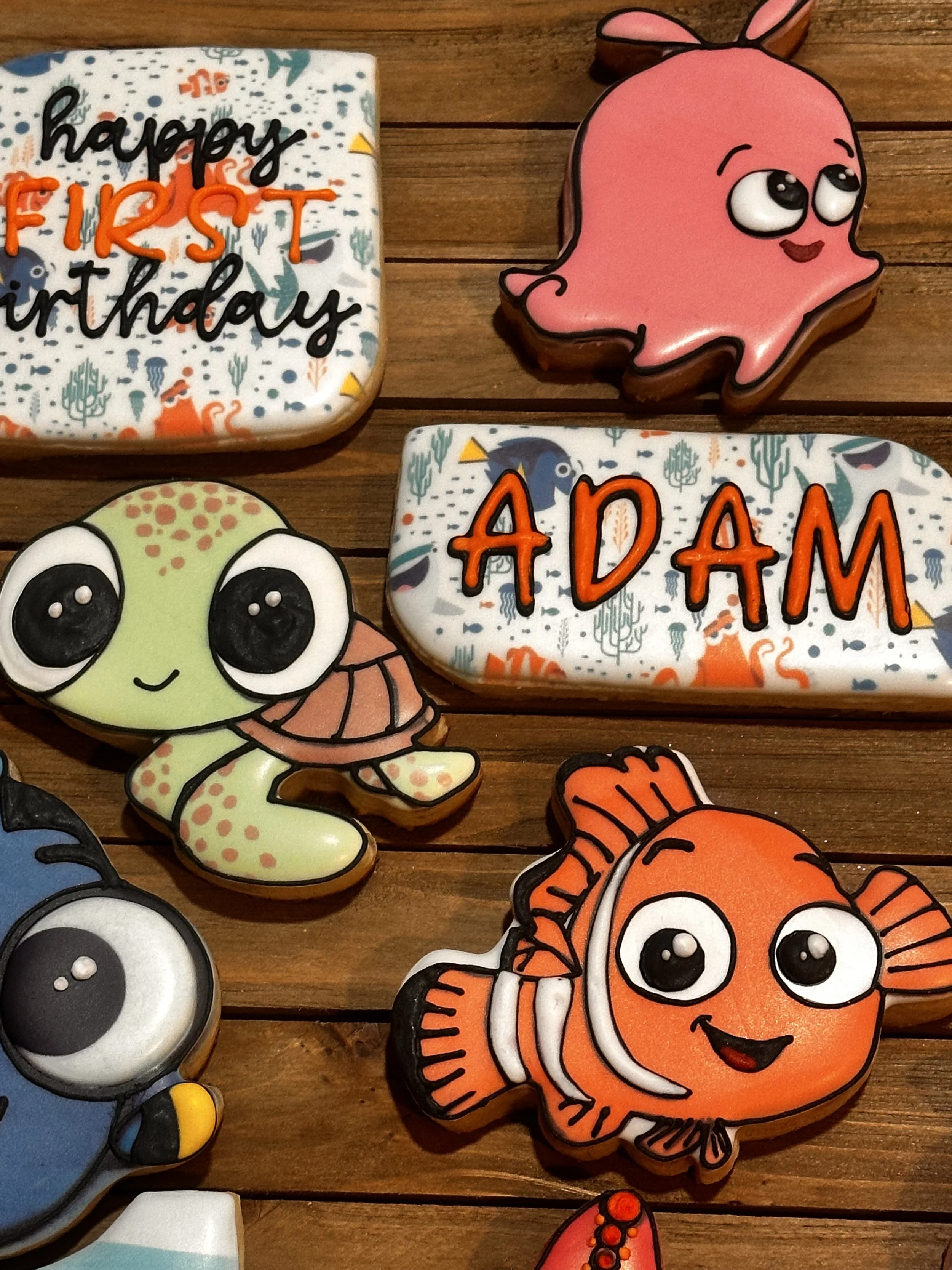 Colorful decorated cookies with ocean and sea creature themes, including a pink ghost, a tortoise, a clownfish, and two rectangular cookies that read 'happy FIRST birthday ADAM'.