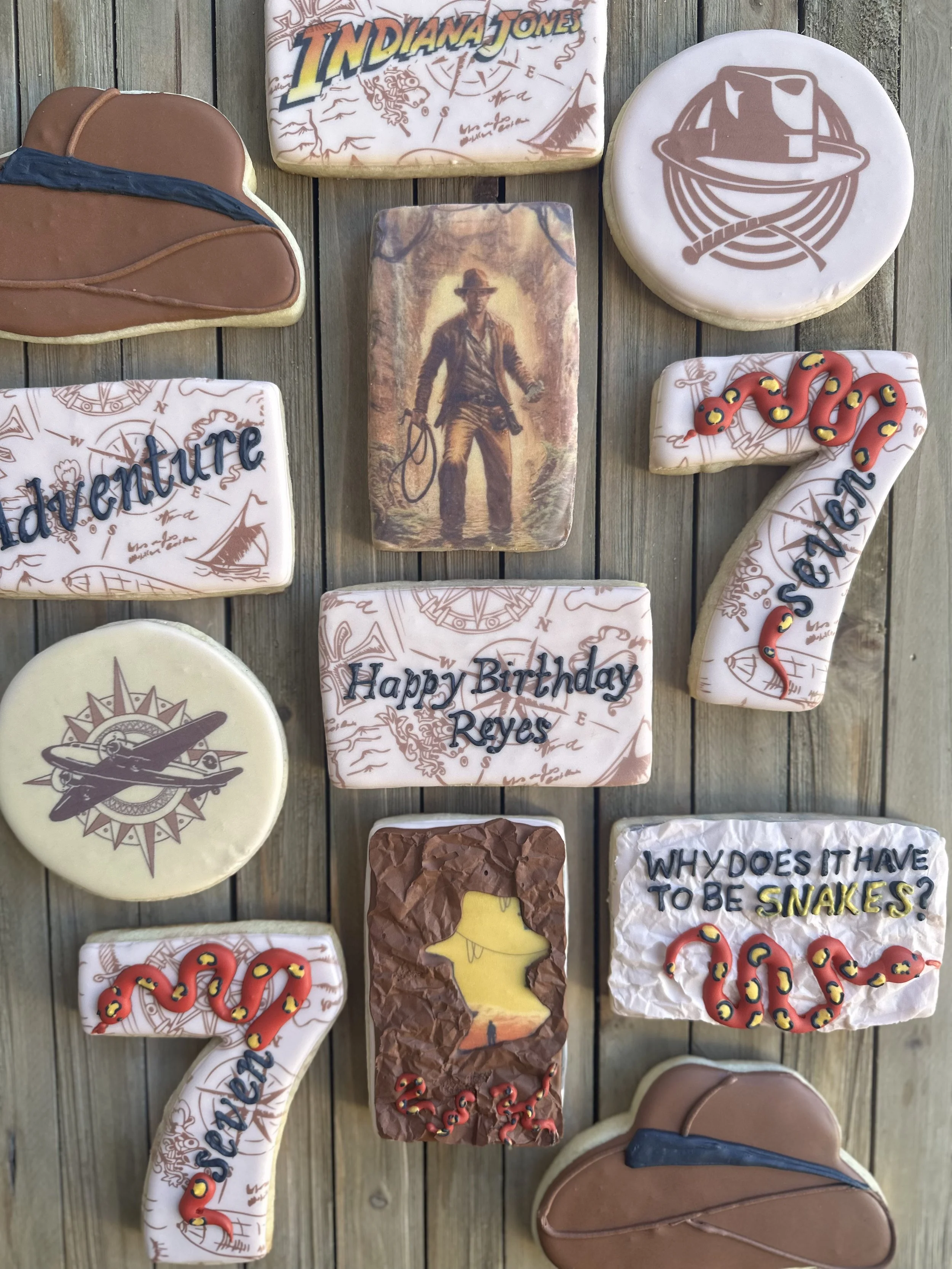 Decorated cookies with adventure and Indiana Jones themes, including a hat, whip, map, snake, and questions about snakes and adventure, on a wooden surface.