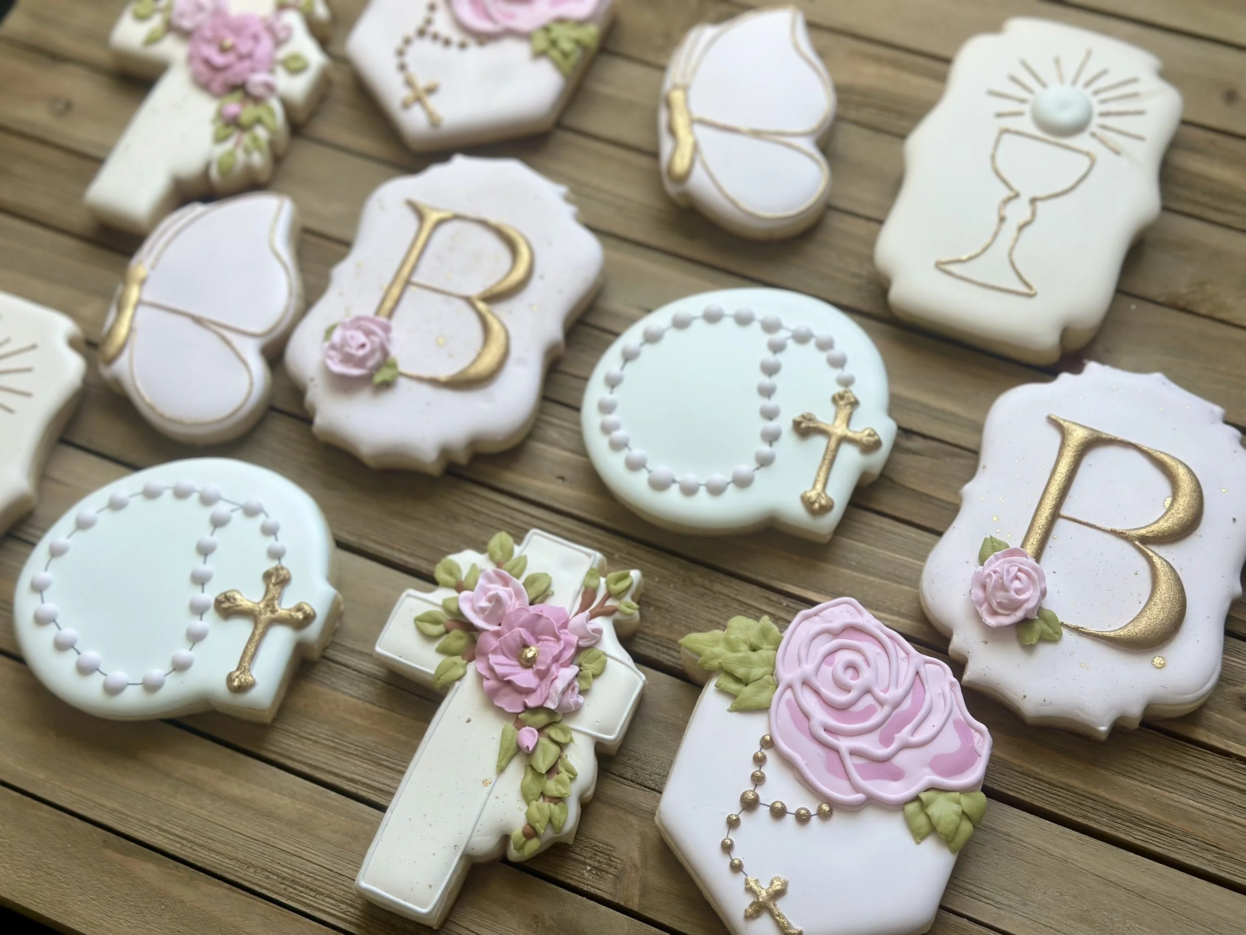 Assorted religious-themed cookies on a wooden surface, decorated with pink and white icing, gold accents, and Christian symbols like crosses, chalices, rosaries, and flowers.