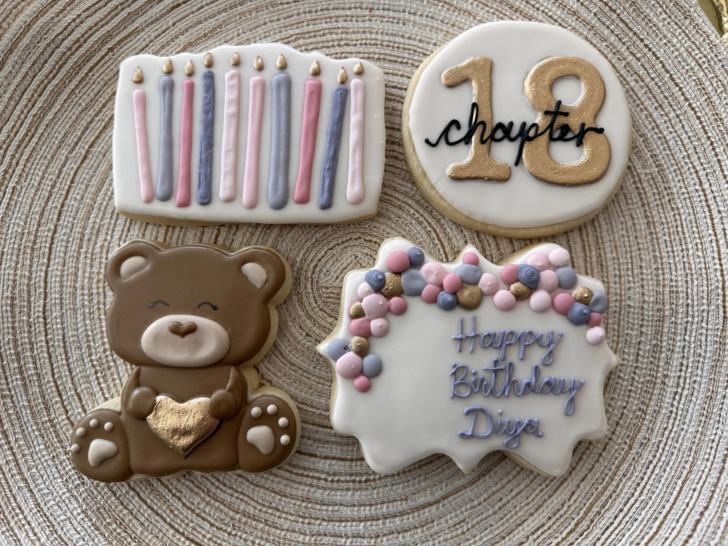 Decorated birthday cookies on a woven round placemat. One cookie has colorful candles, another says '10 chapter', a third is a cute teddy bear holding a gold heart, and the fourth has the message 'Happy Birthday Diya' with pink, gray, and gold dots.