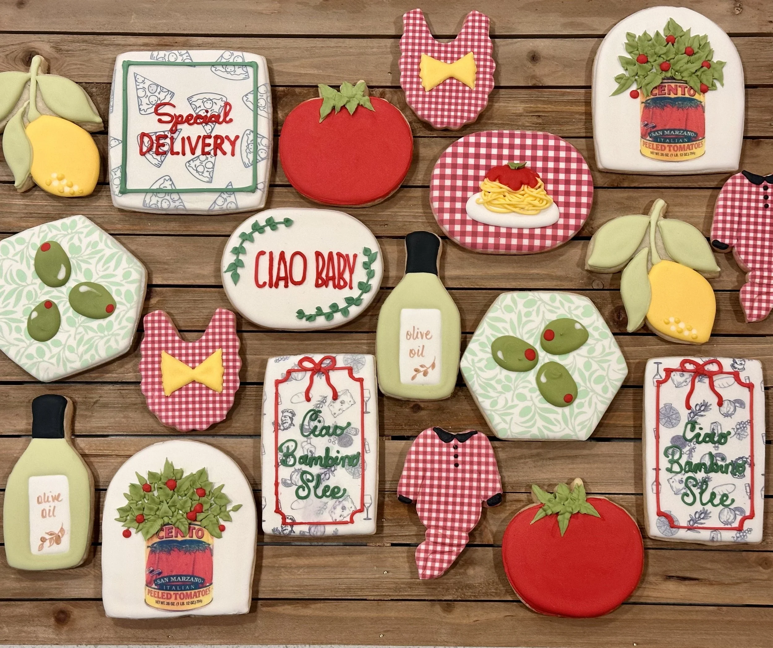 Assorted decorated cookies on a wooden surface, themed around Italian cuisine and groceries, including tomatoes, olives, pasta, garlic, oregano, and pizza, with some cookies featuring phrases like "Special Delivery," "Ciao Baby," and "Ciao Bambino Sl