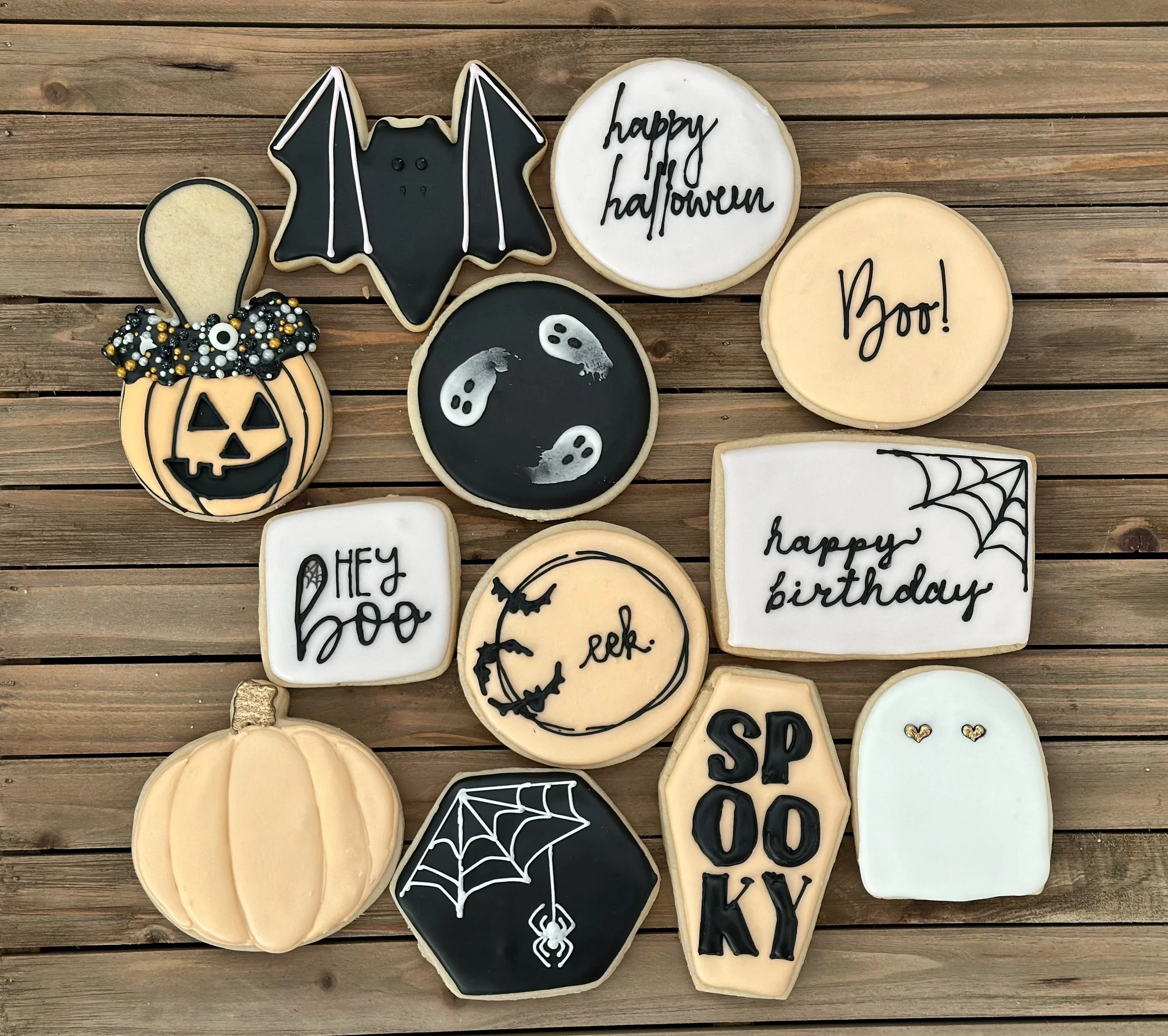 Assorted Halloween-themed cookies on a wooden surface, including a bat, pumpkin, ghost, spiderweb, and individual words like "Boo!", "happy halloween", "happy birthday", and "spooky".