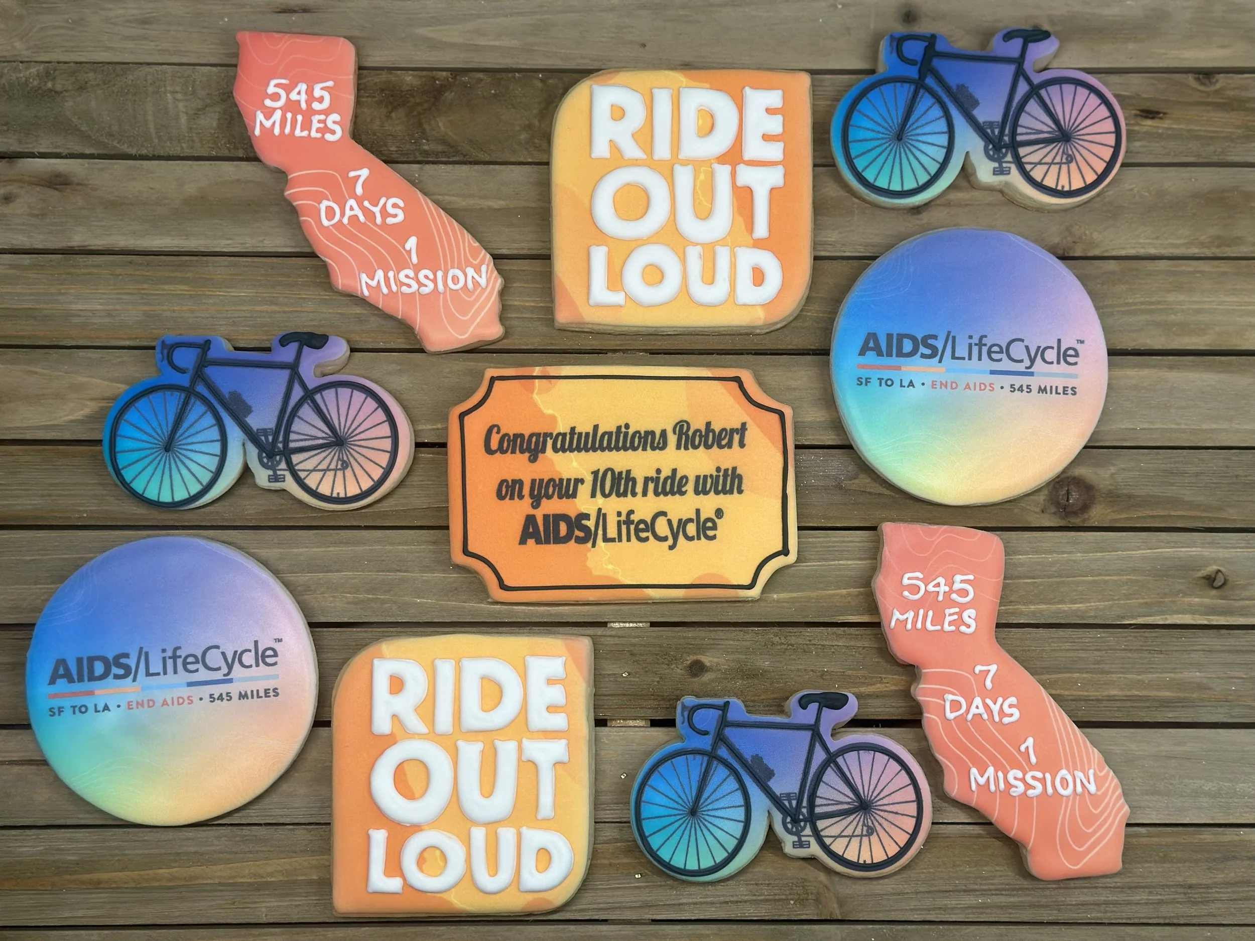 Collection of badges and signs celebrating a 10th bike ride for AIDS/LifeCycle on a wooden surface, including messages like 'Ride Out Loud,' 'Congratulations Robert on your 10th ride with AIDS/LifeCycle,' and details about the event such as '545 mile