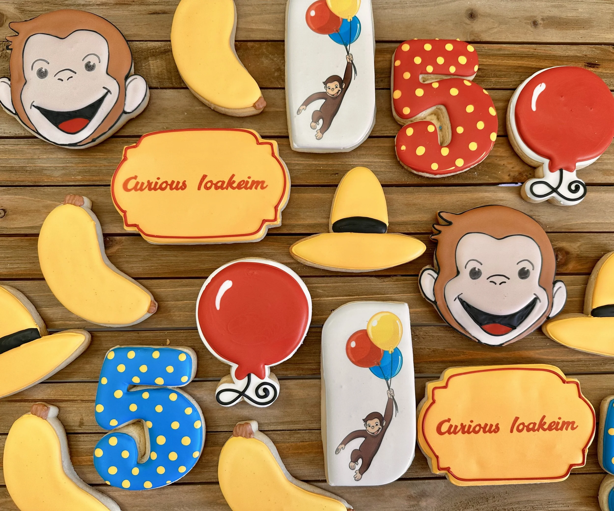 Decorated cookies with monkey theme, featuring monkey face, bananas, balloons, and the phrase 'Curious Loakeim' on orange and yellow backgrounds, arranged on a wooden surface.