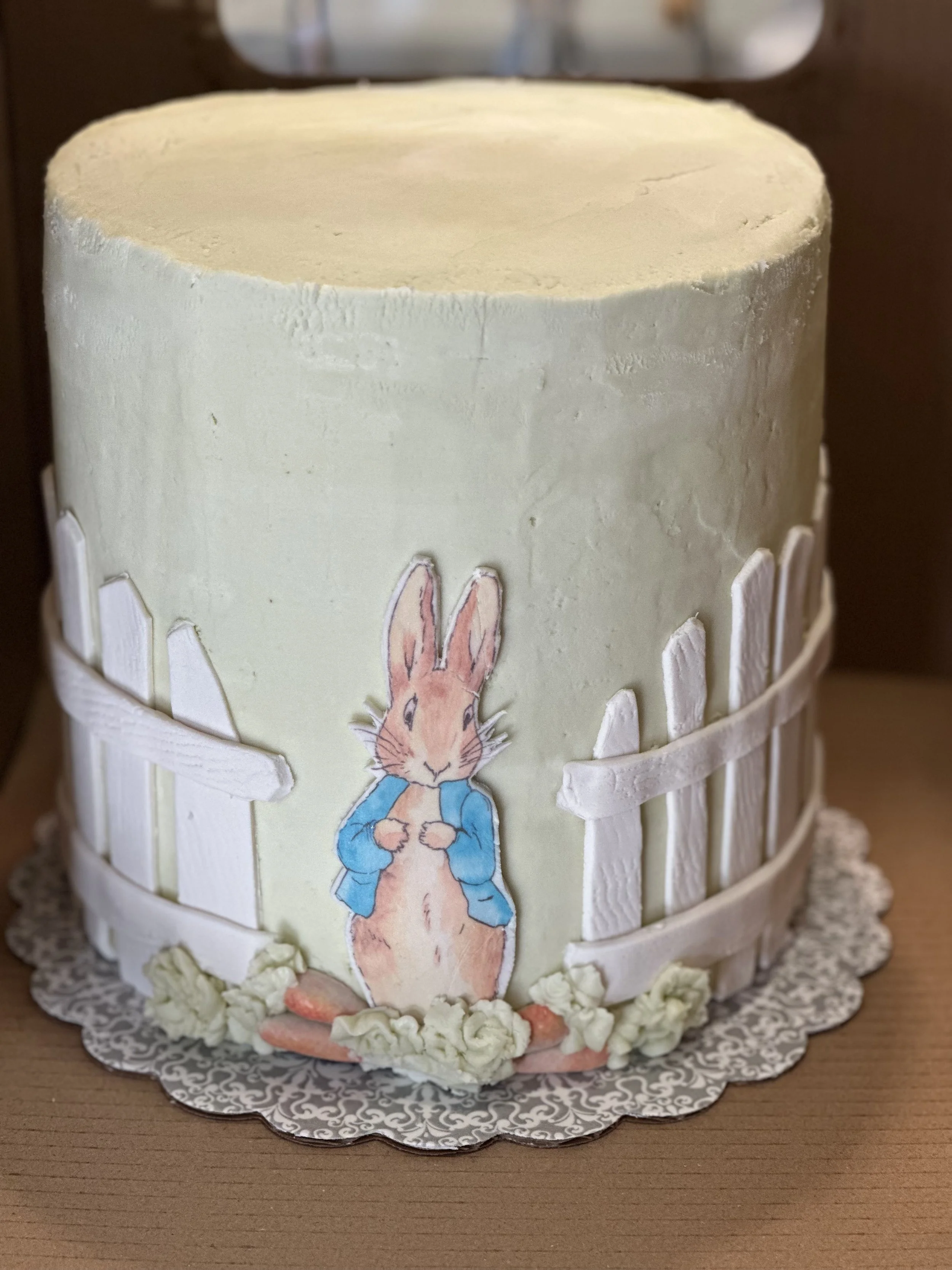Easter cake with white frosting, decorative white picket fence, paper bunny illustration wearing a blue jacket, and small white flowers at the base on a patterned silver cake board.