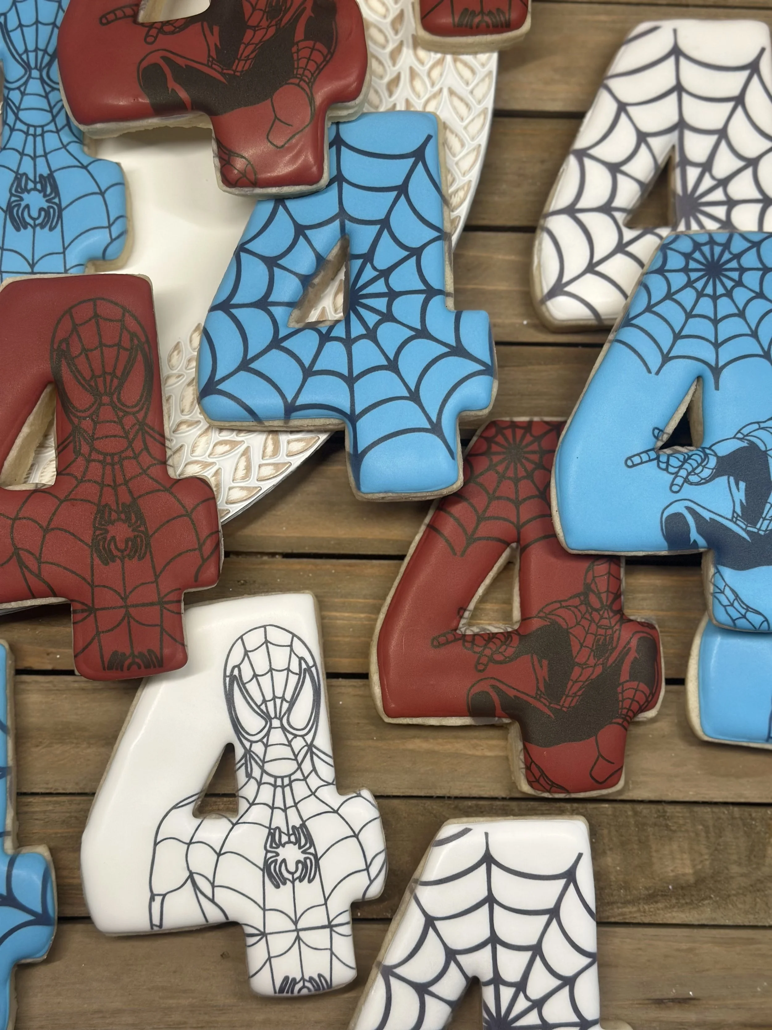 Cookies shaped like the number four, decorated with Spider-Man and spider web designs in red, blue, and white icing on a wooden surface.