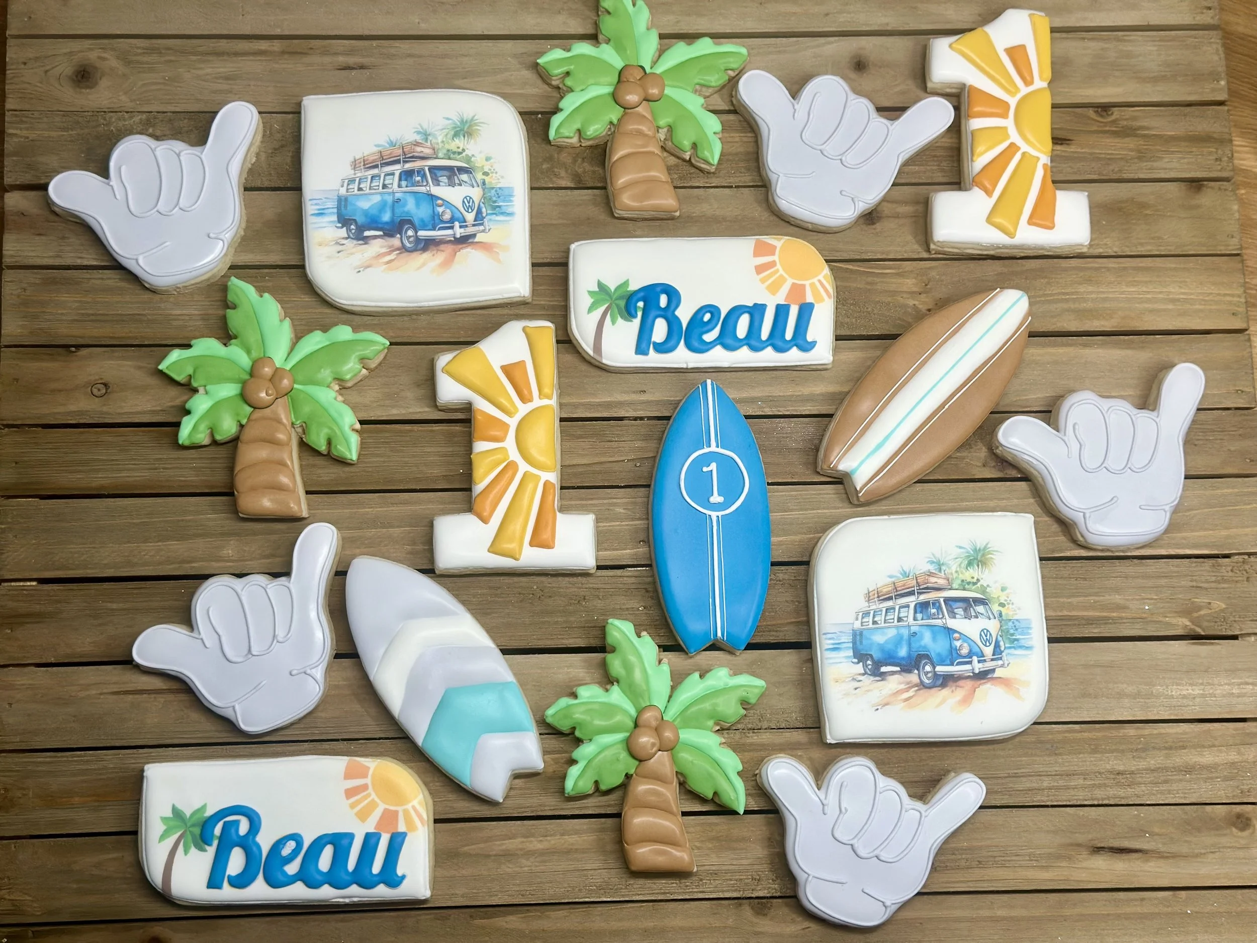 Assorted colorful decorated cookies with beach and tropical themes, including palm trees, surfboards, buses, the word "Beau," and hand signs, arranged on a wooden surface.