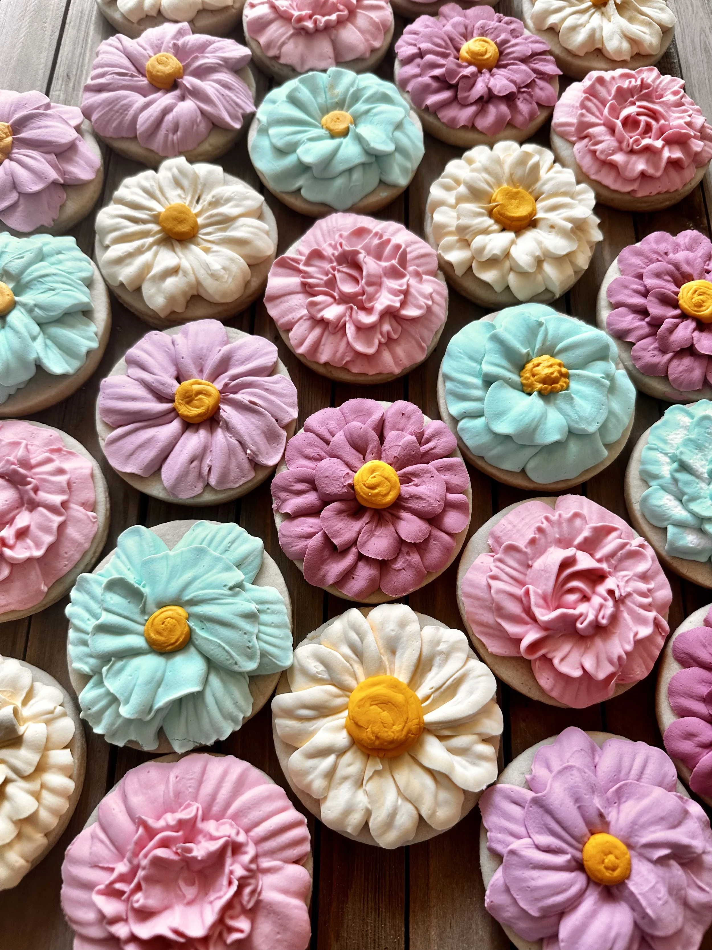 Assorted decorated sugar cookies shaped like flowers in pastel colors with ruffled icing petals and yellow centers, arranged on a wooden surface.