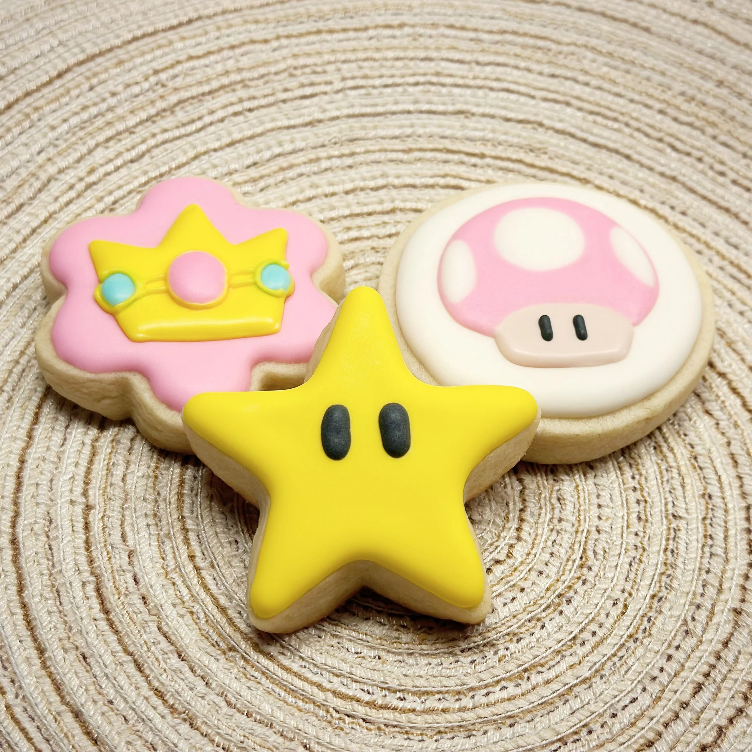 Three decorated cookies on a woven circular mat: a pink flower-shaped cookie with a yellow crown design, a white round cookie with a pink mushroom design, and a yellow star-shaped cookie with black eyes.