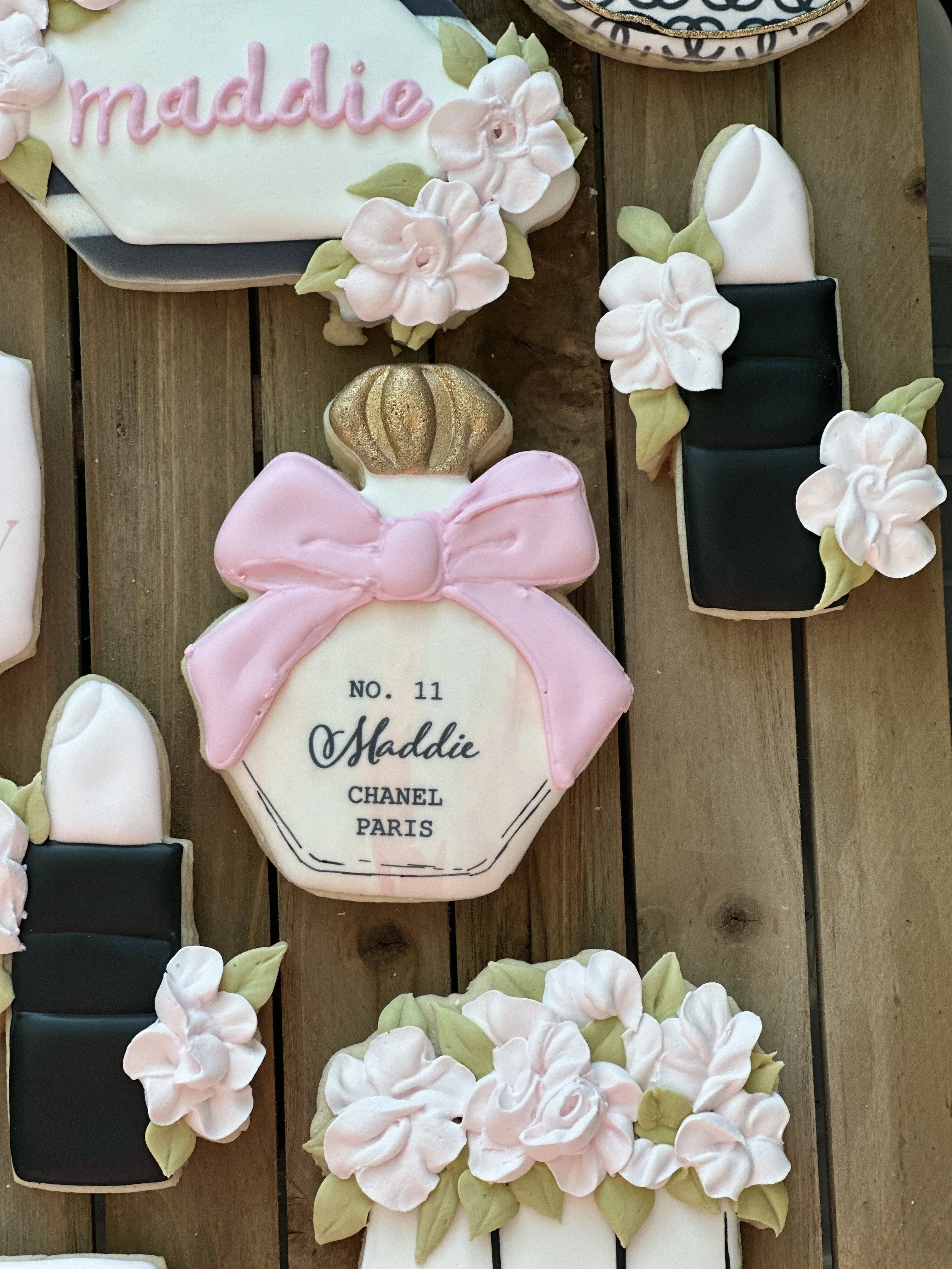 Decorative cookies designed to look like a perfume bottle and various flowering arrangements, with a pink and white color scheme, on a wooden surface.