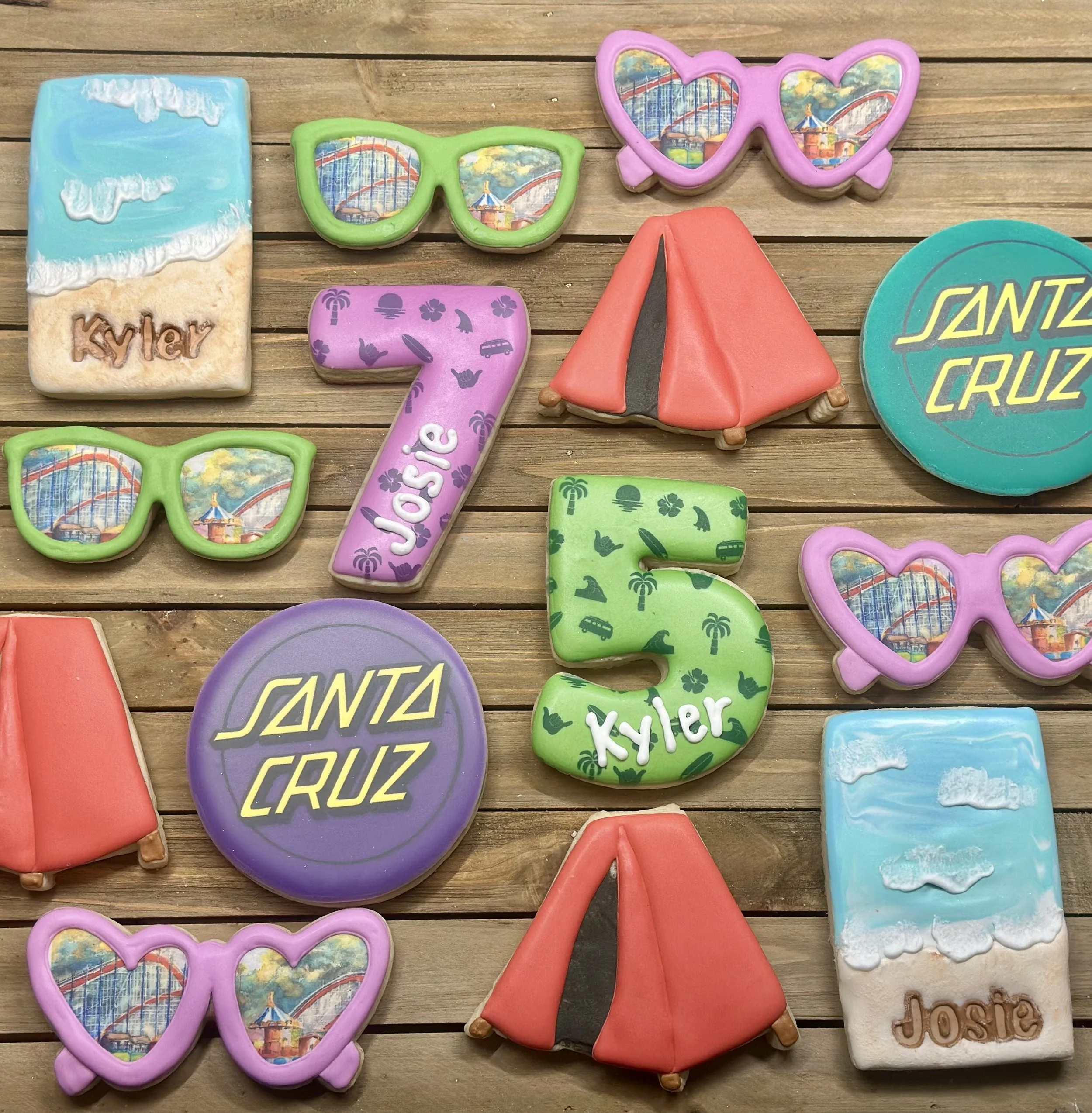 Colorful decorated cookies shaped like sunglasses, a tent, the number 7 with Josie written, the number 5 with Tyler written, and circular cookies with Santa Cruz logos, all arranged on a wooden surface.